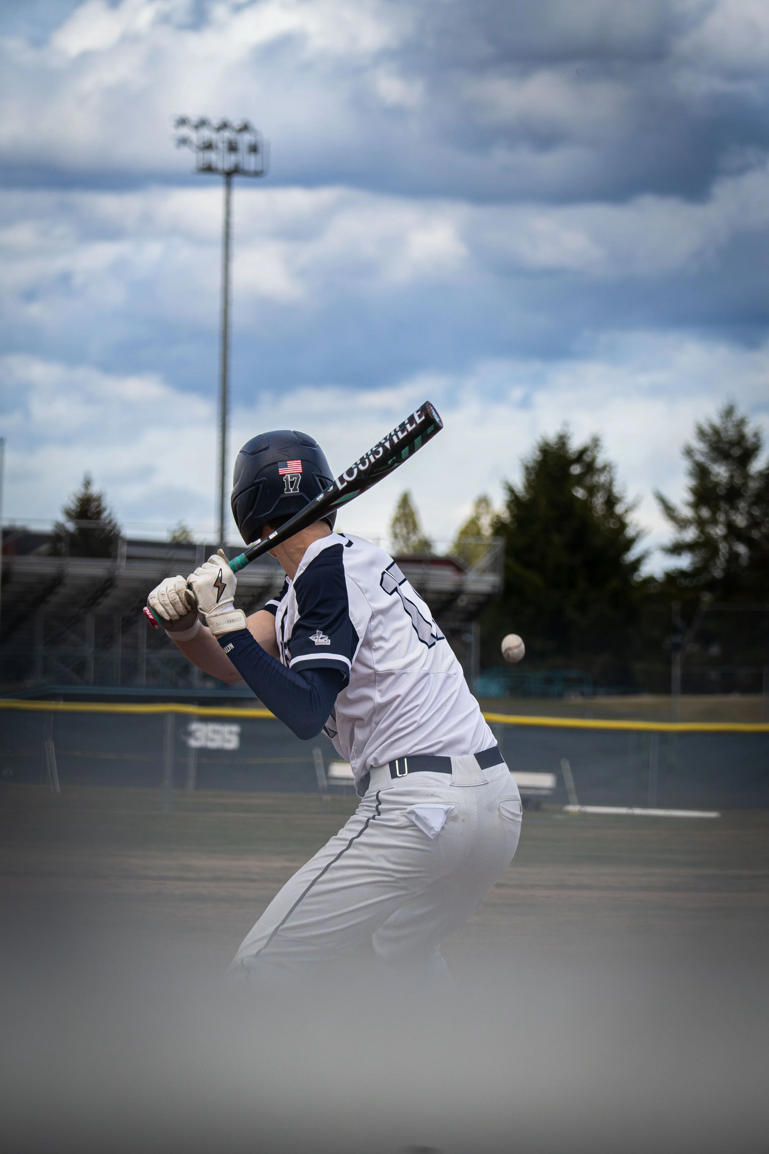 A baseball player in a white uniform with dark blue detailing, wearing a helmet with an American flag and number 17, swings a bat on a baseball field. Overcast sky with dark clouds is visible in the background.