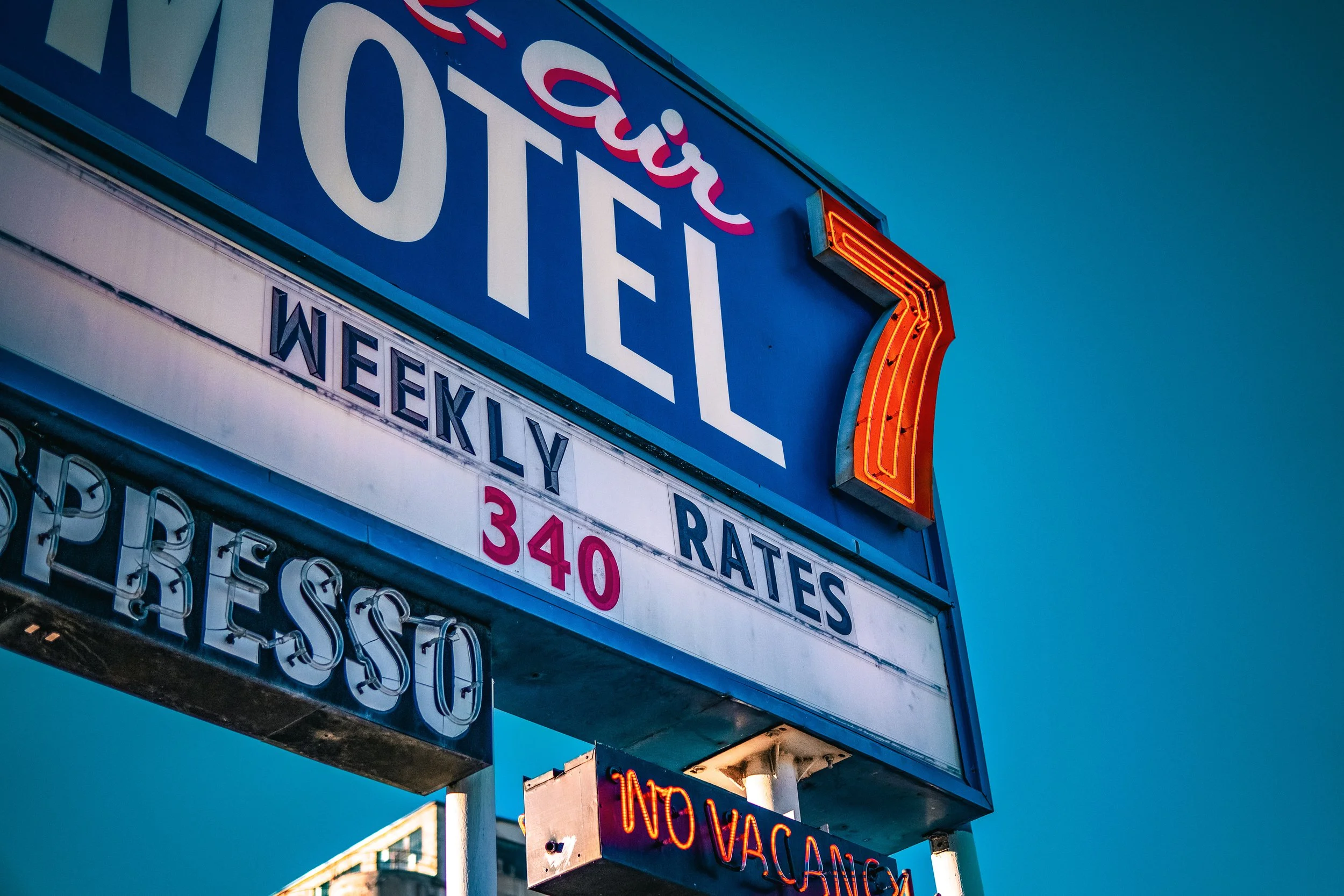 Vintage motel sign with weekly rates, price shown as 340, neon lights, and "No Vacancy" sign, against blue sky.