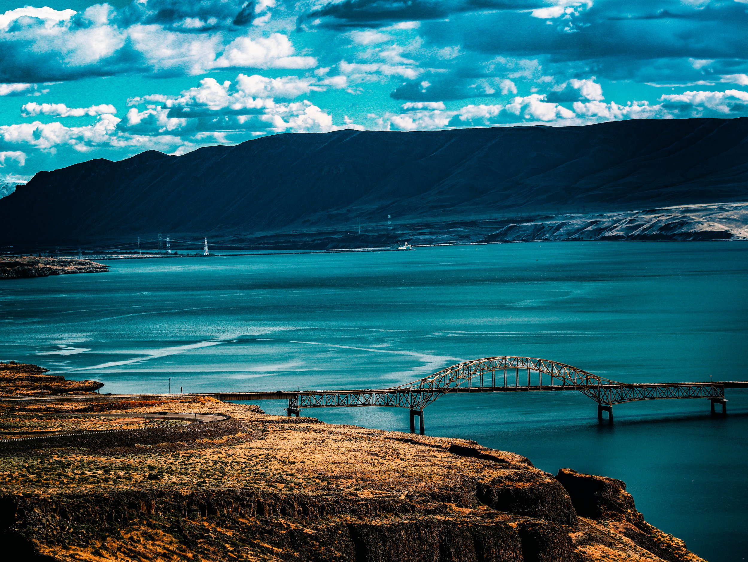 A landscape of a large body of water with a bridge extending across it, set against a backdrop of mountains under a partly cloudy sky.