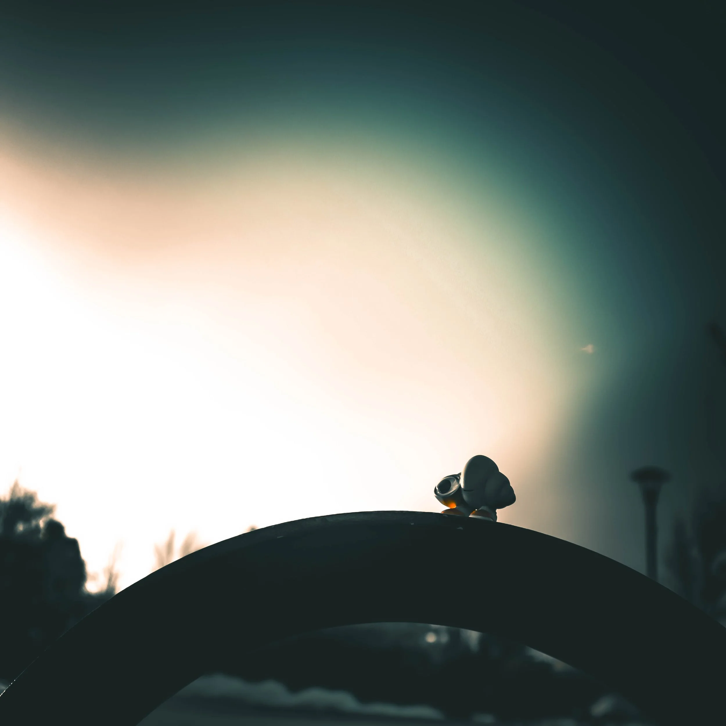 A small marcel the shell figure on a bench with a blurred outdoor scene in the background, including trees and sky, during sunset or sunrise.