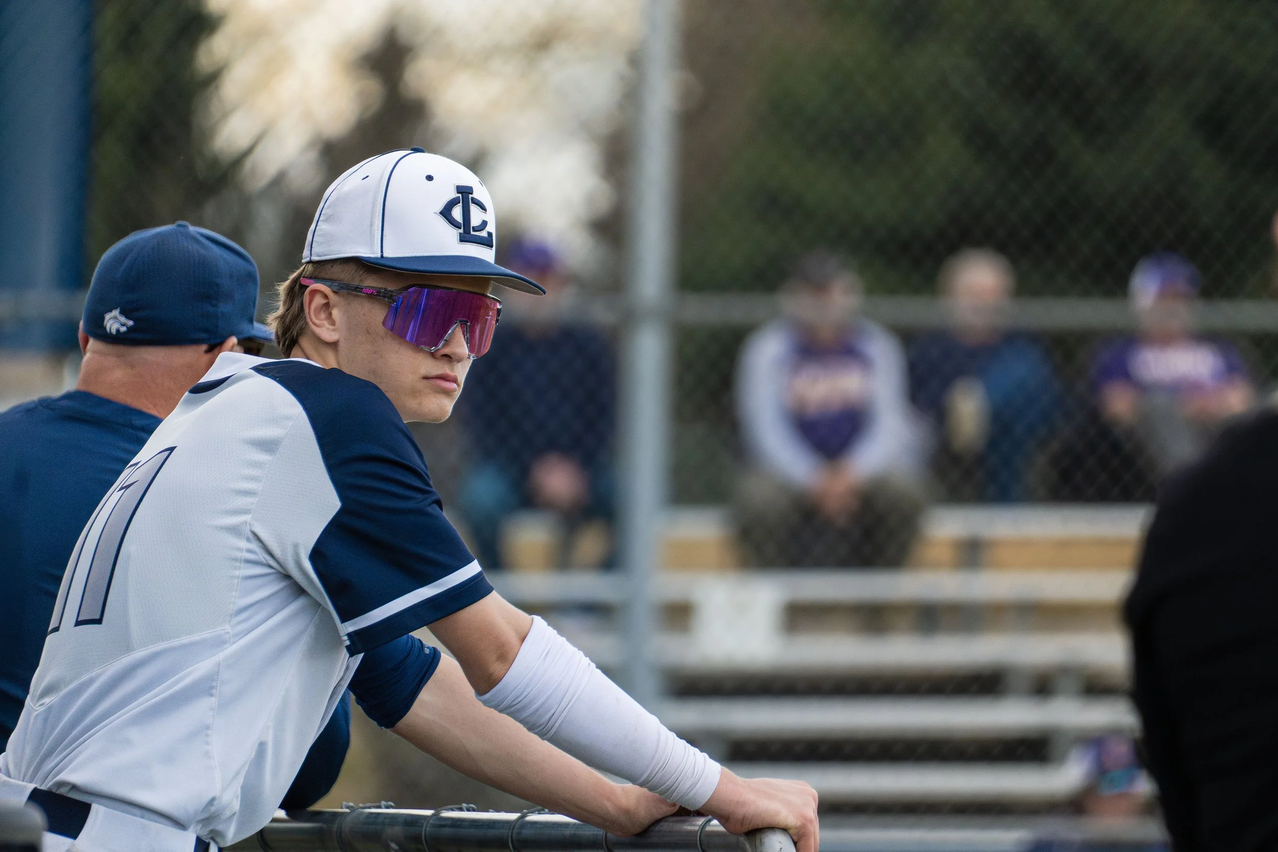Baseball player wearing sunglasses and cap, standing near the dugout with spectators in the background.