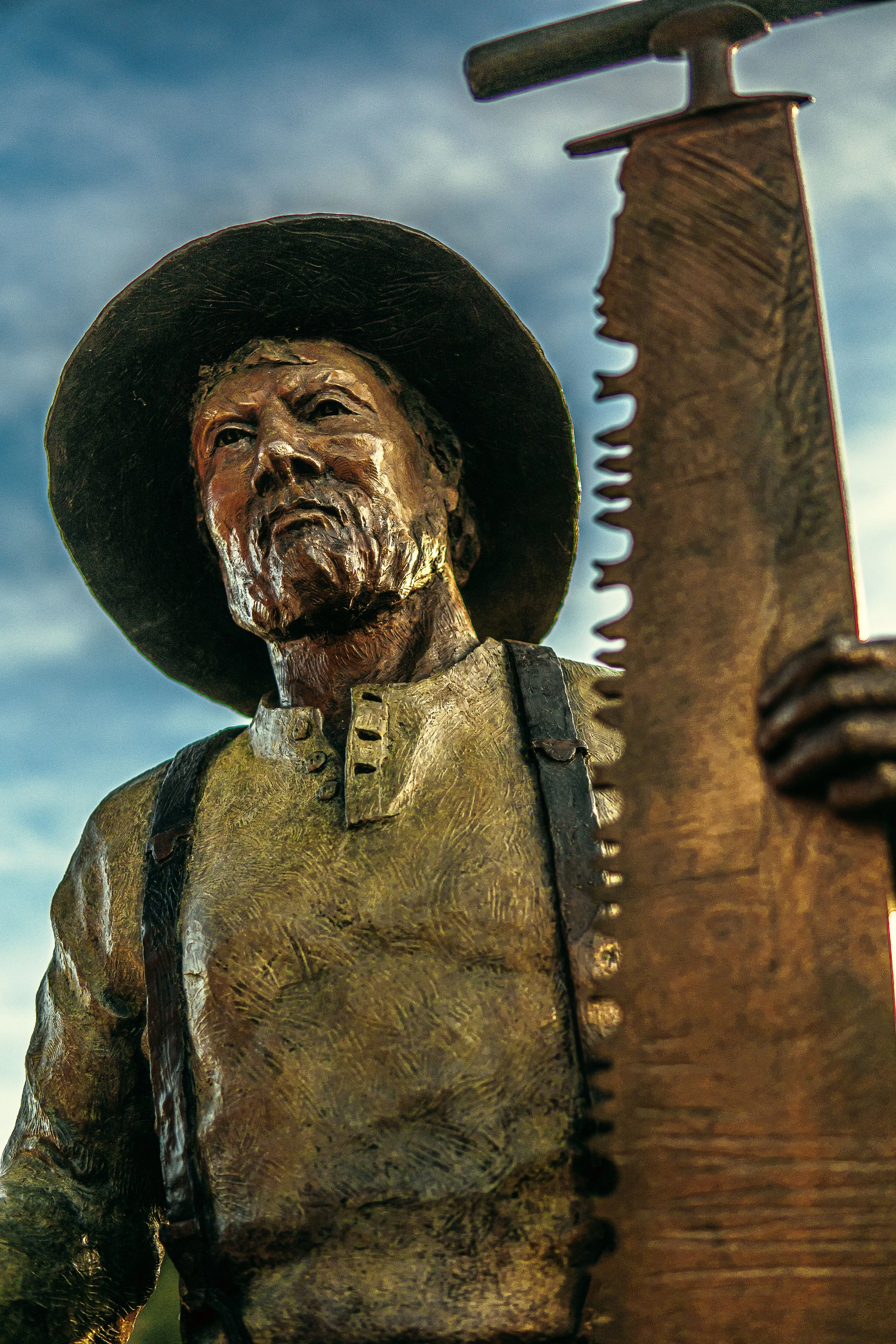 Bronze statue of a man holding a large saw, wearing a hat, with a blue sky background.
