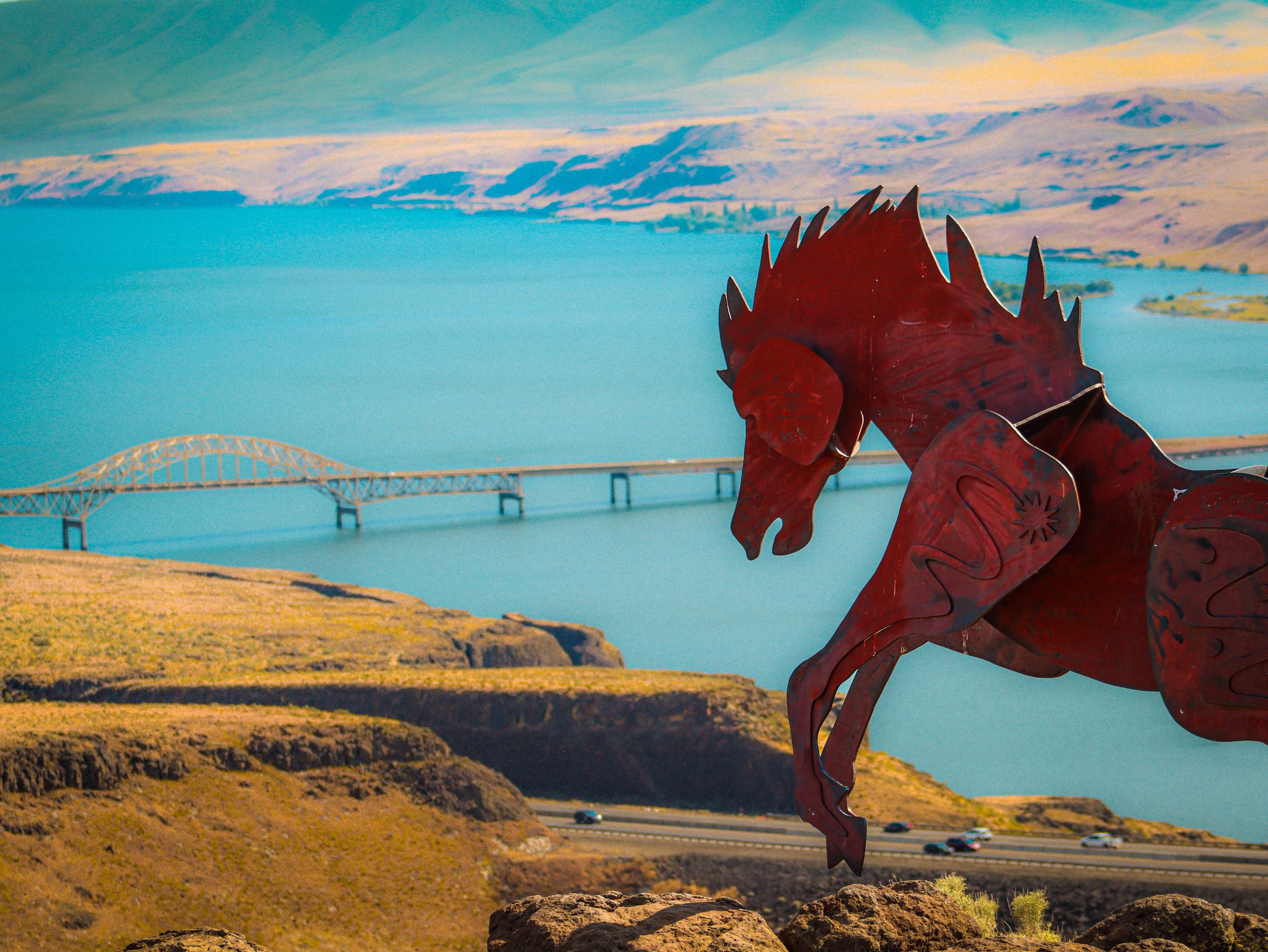 Red metal sculpture of a horse with a mountainous landscape, bridge, and body of water in the background.