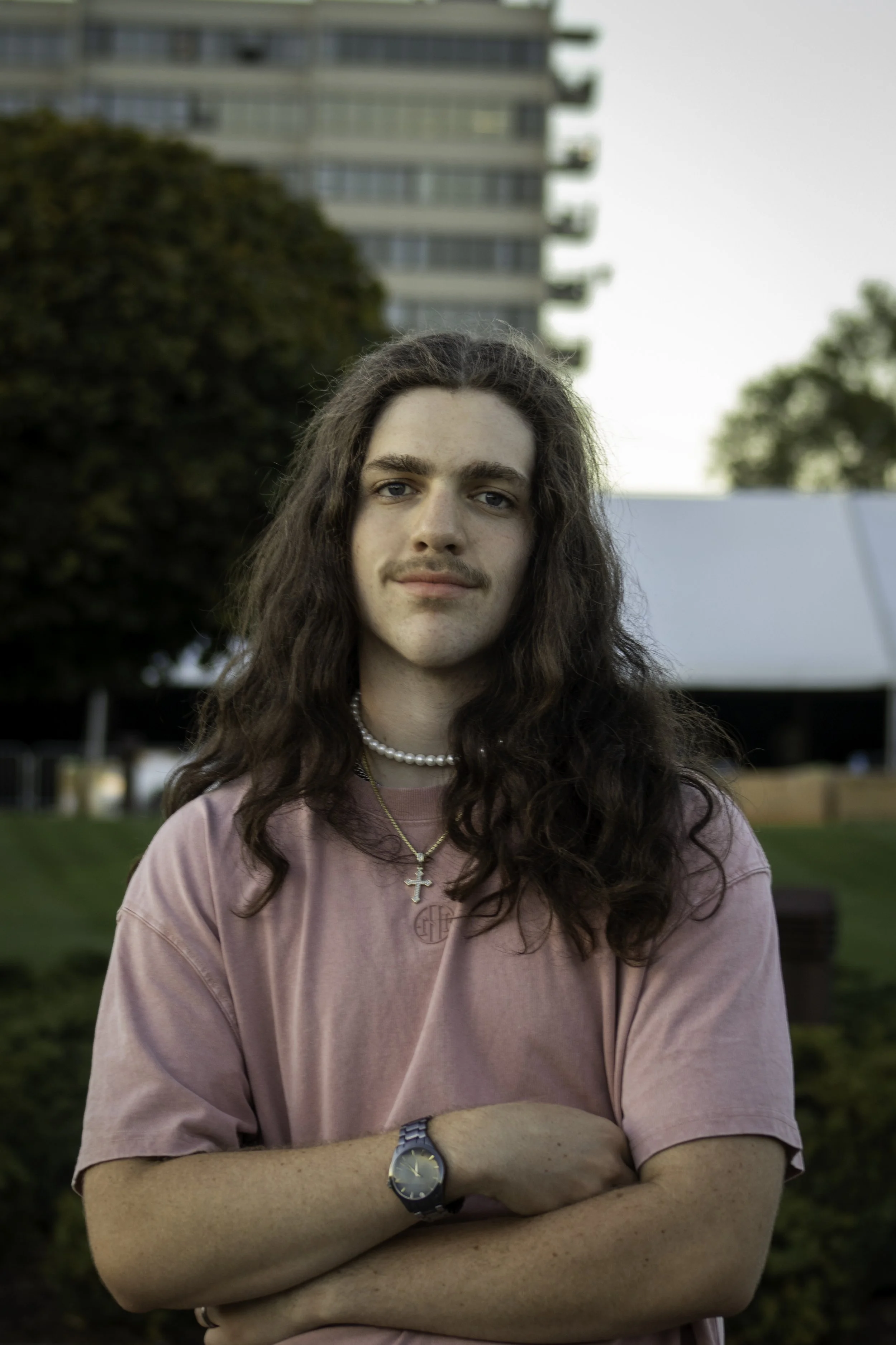 Person with long hair and mustache, wearing a pink shirt, pearl necklace, and cross pendant, standing outdoors with a building and trees in the background.