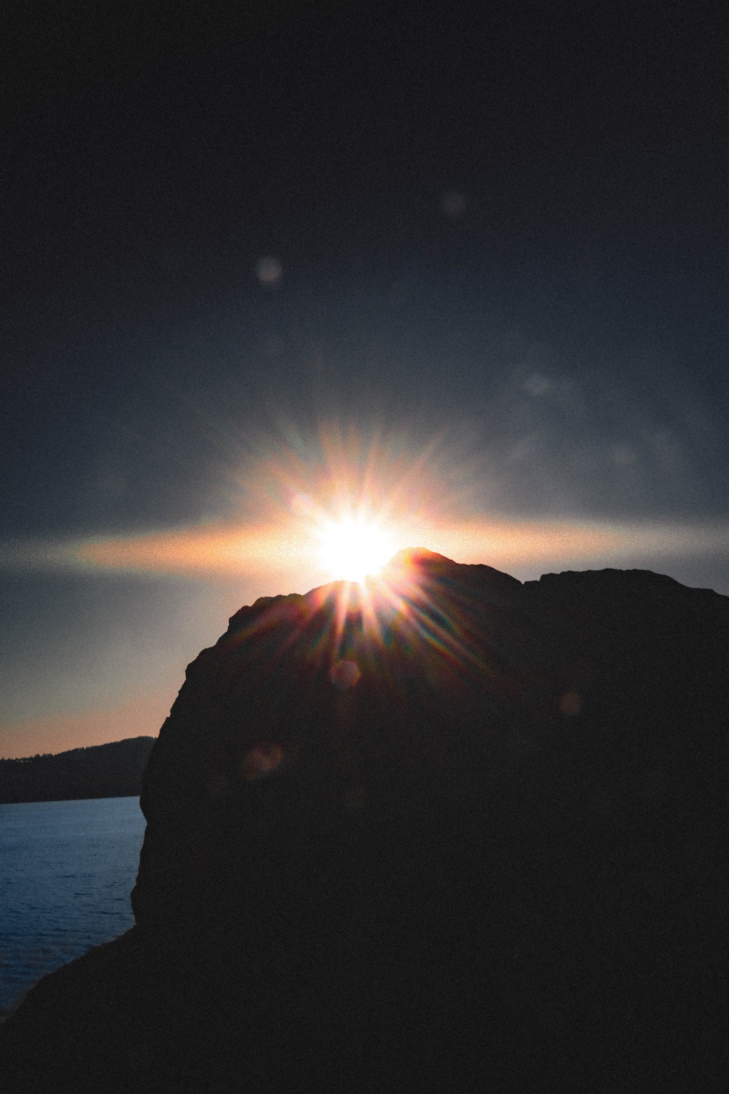 Sun setting behind a rocky cliff over a body of water, creating a starburst effect with rays of light.