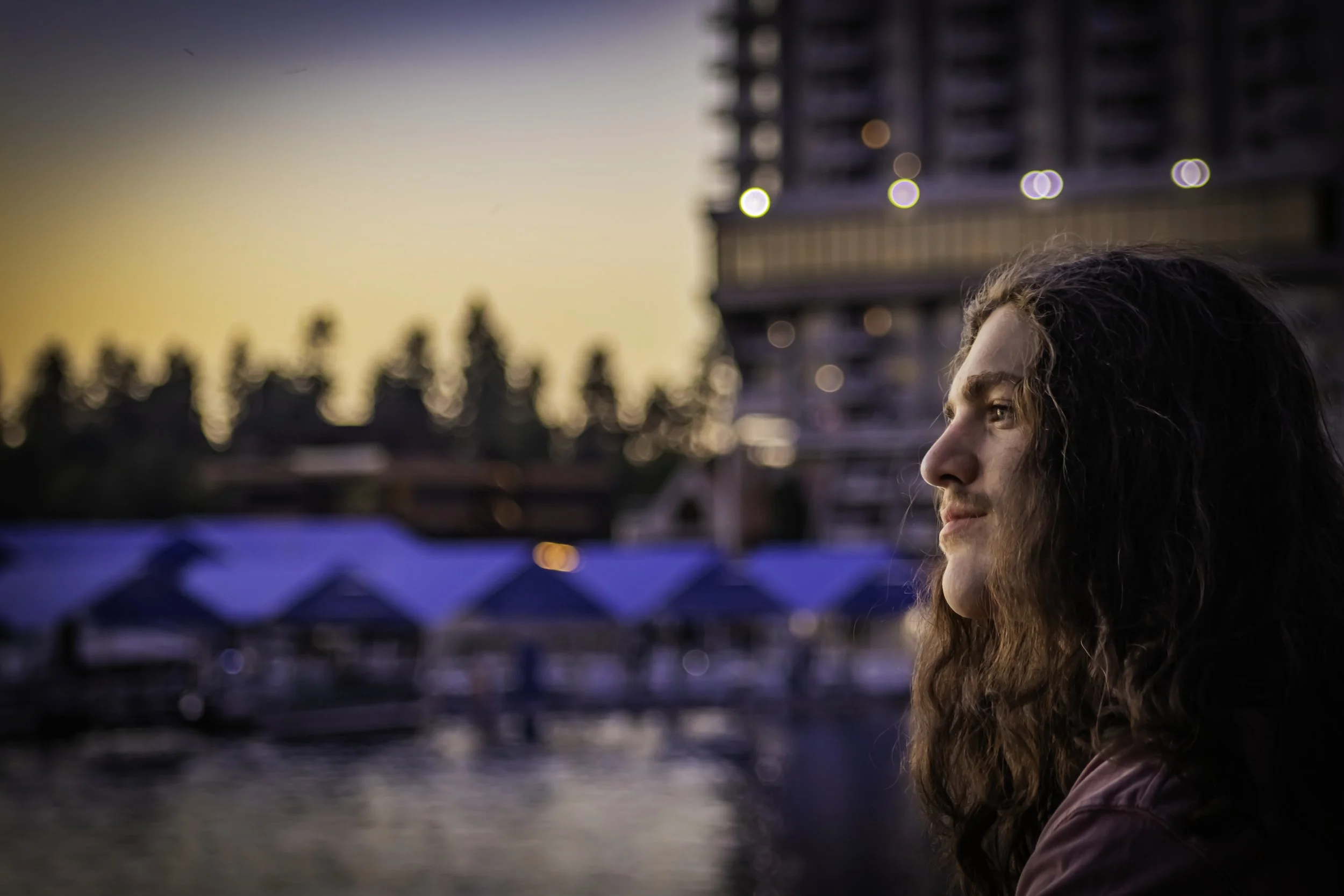 Man with long hair and mustache gazing, urban waterfront background, sunset lighting.