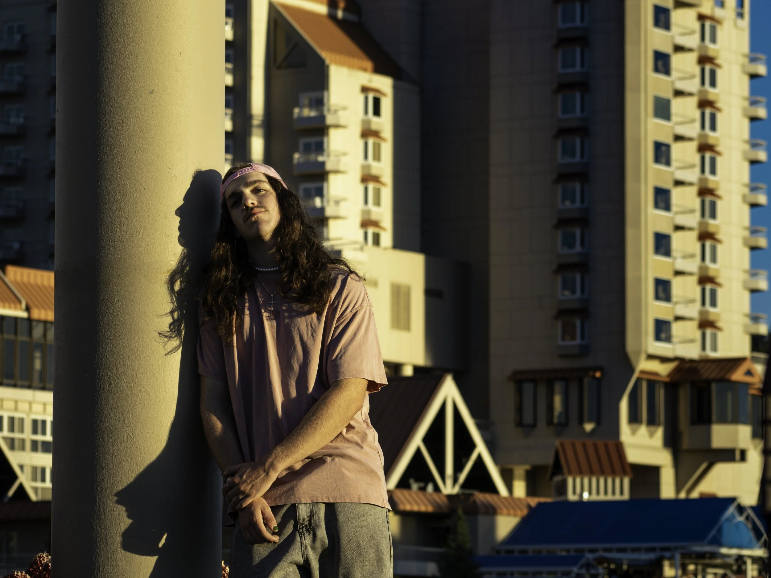 Man with long hair leaning casually against a column in urban setting, wearing a pink shirt and cap, with buildings in the background.
