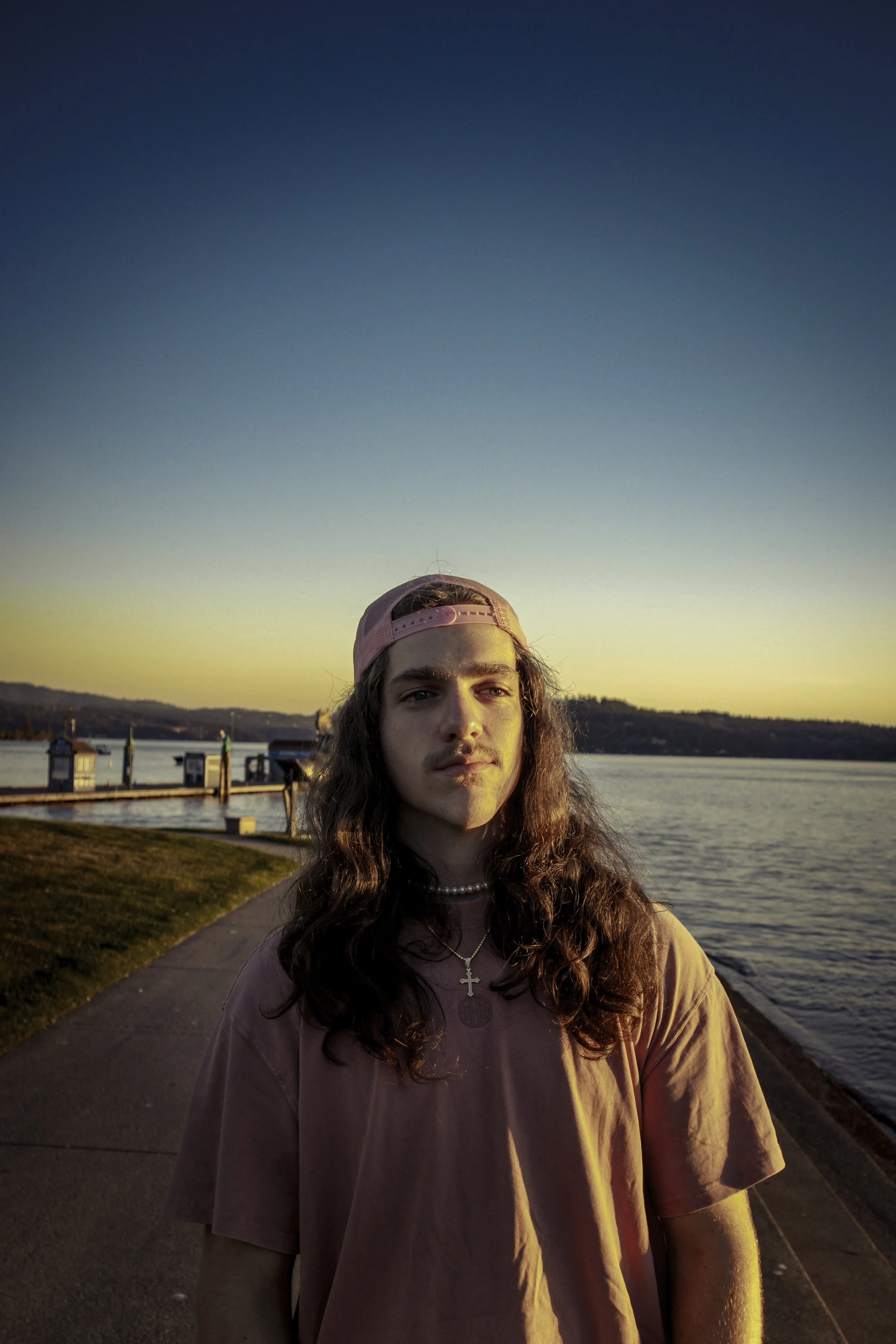 A person with long hair and a pink cap stands by a waterfront at sunset. They wear a pink shirt and pearl necklace with a cross pendant. The background features a calm body of water and distant hills under a clear sky.