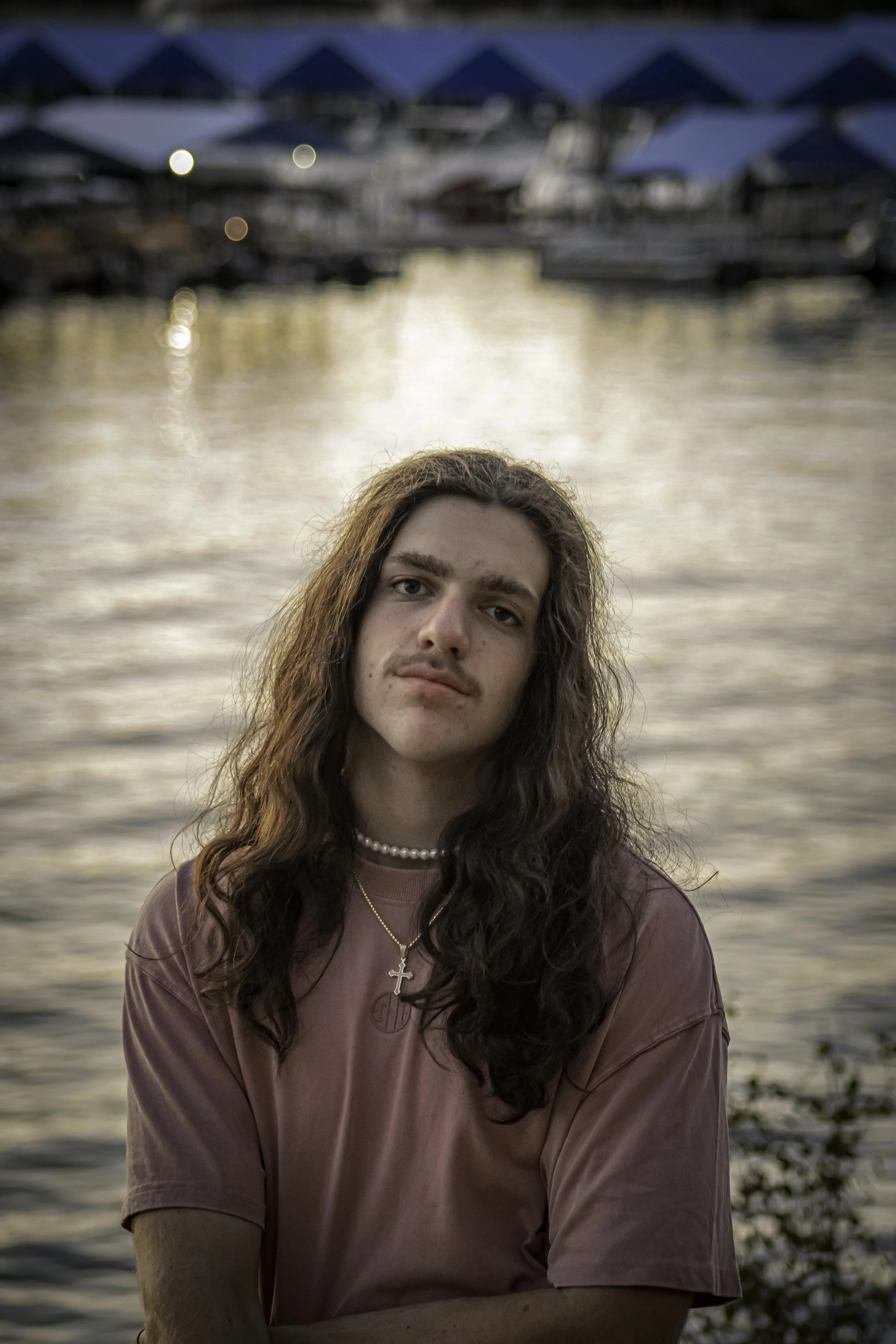 Person with long hair and a mustache near a body of water, wearing a pink shirt and necklaces, with boats and dock structures in the background at sunset.
