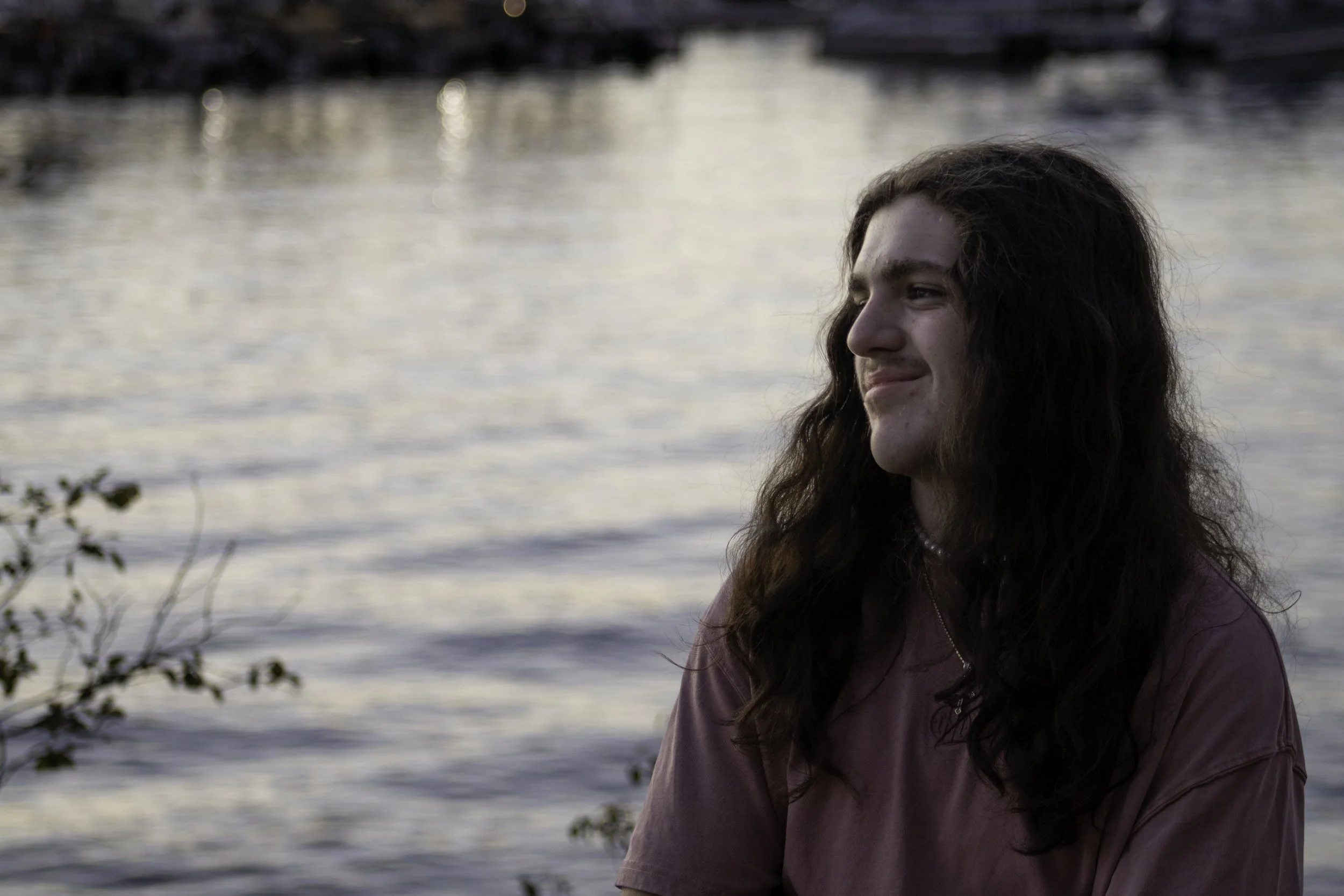 Person with long hair smiling by a body of water, wearing a pink shirt, with trees in the foreground.