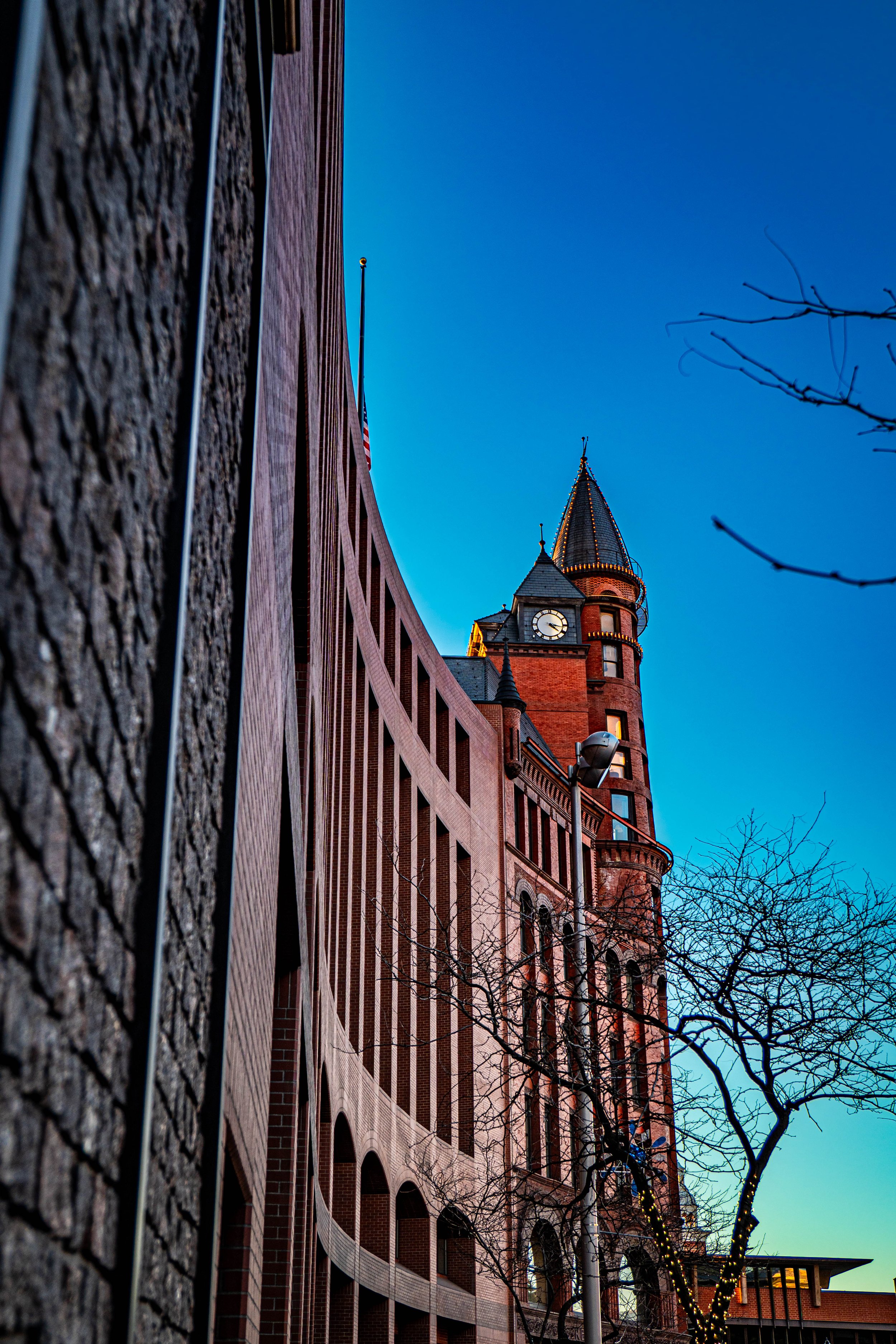 Low-angle view of a historic brick building with a clock tower and a pointed turret, during twilight with a clear blue sky, leafless trees, and decorative string lights.