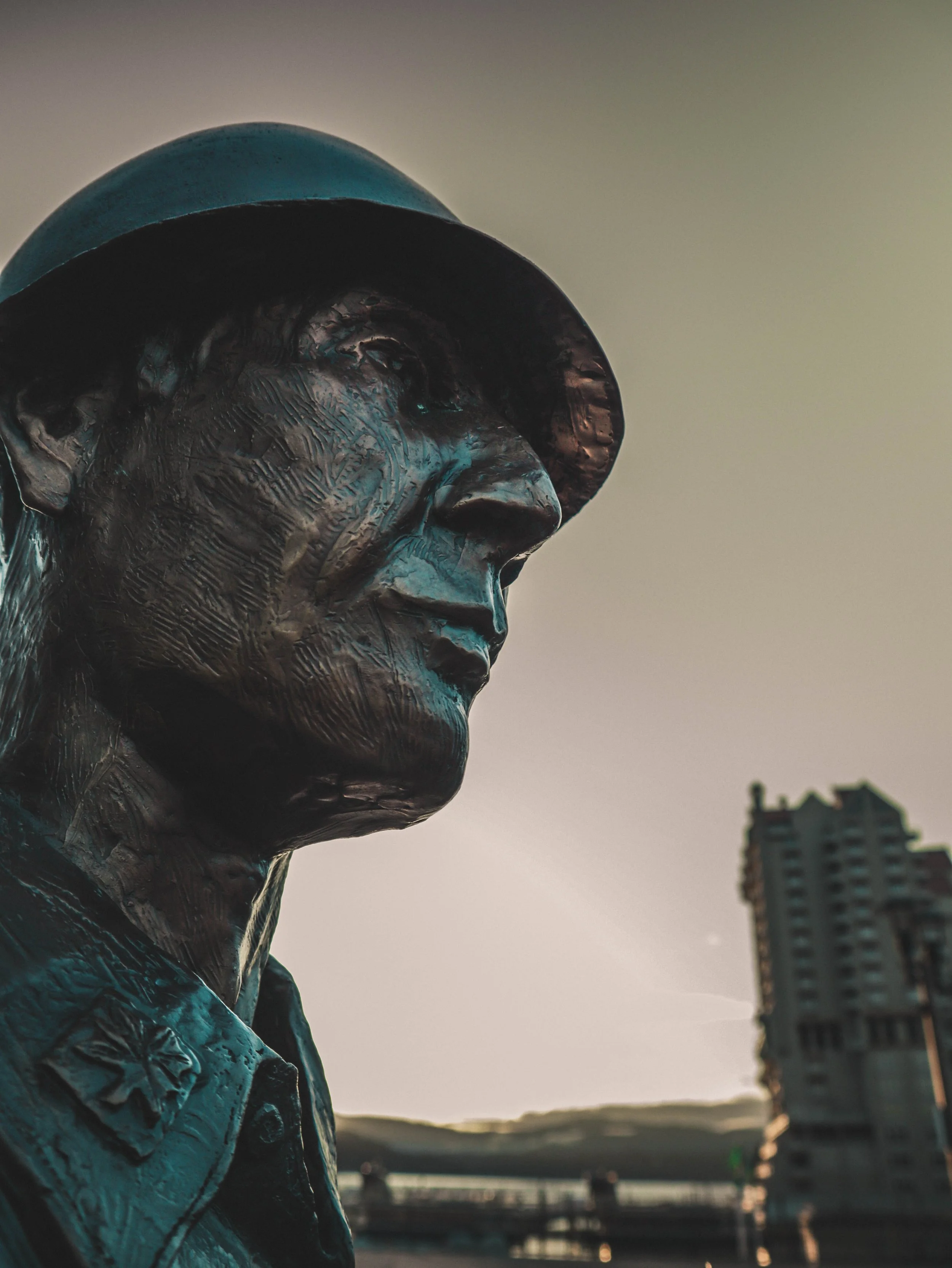 Close-up of a bronze bust of a man wearing a hat, with a cityscape and river in the background during sunset.