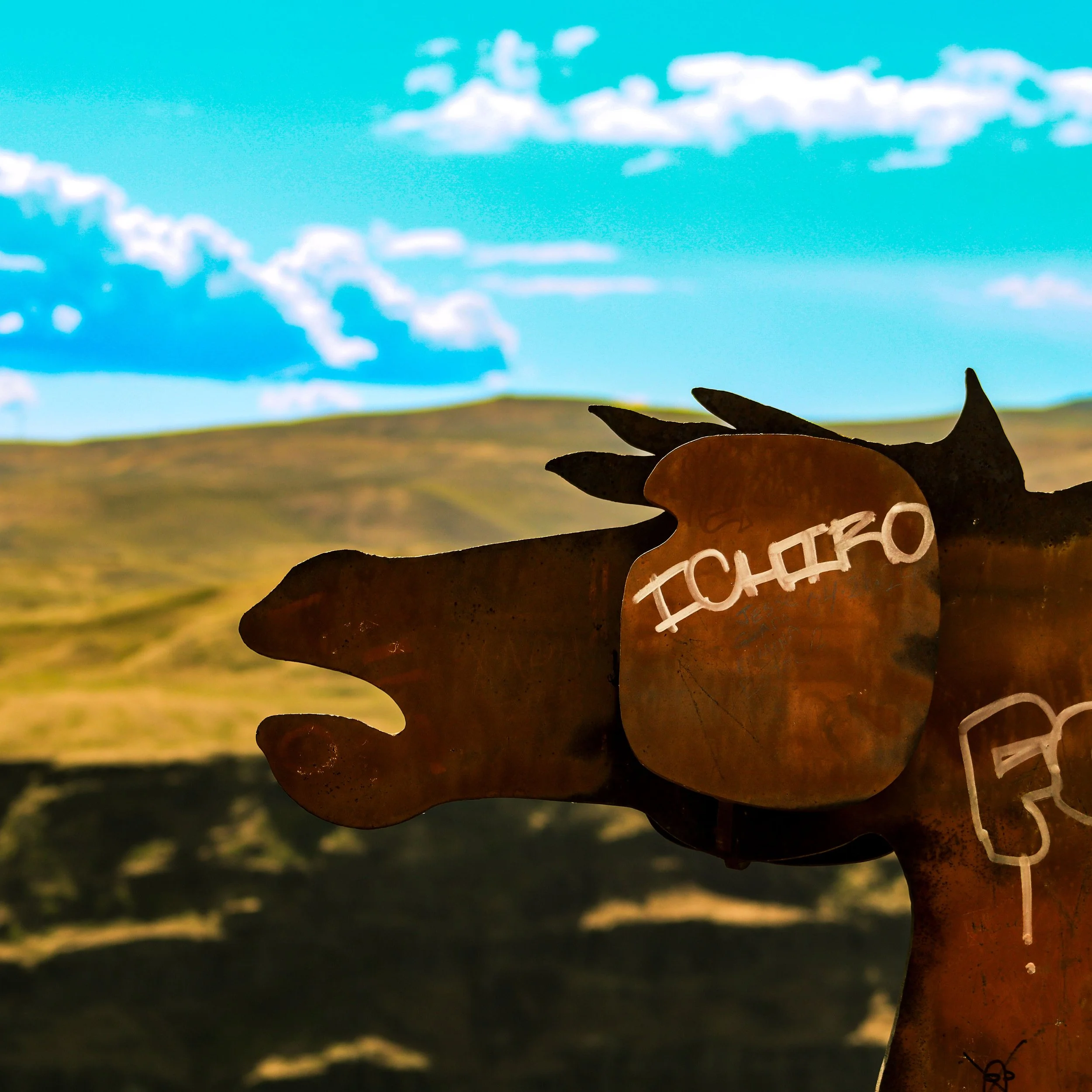 Close-up silhouette of a metal cutout sign with the word 'pachinko' against a bright blue sky with scattered clouds and a landscape in the background.