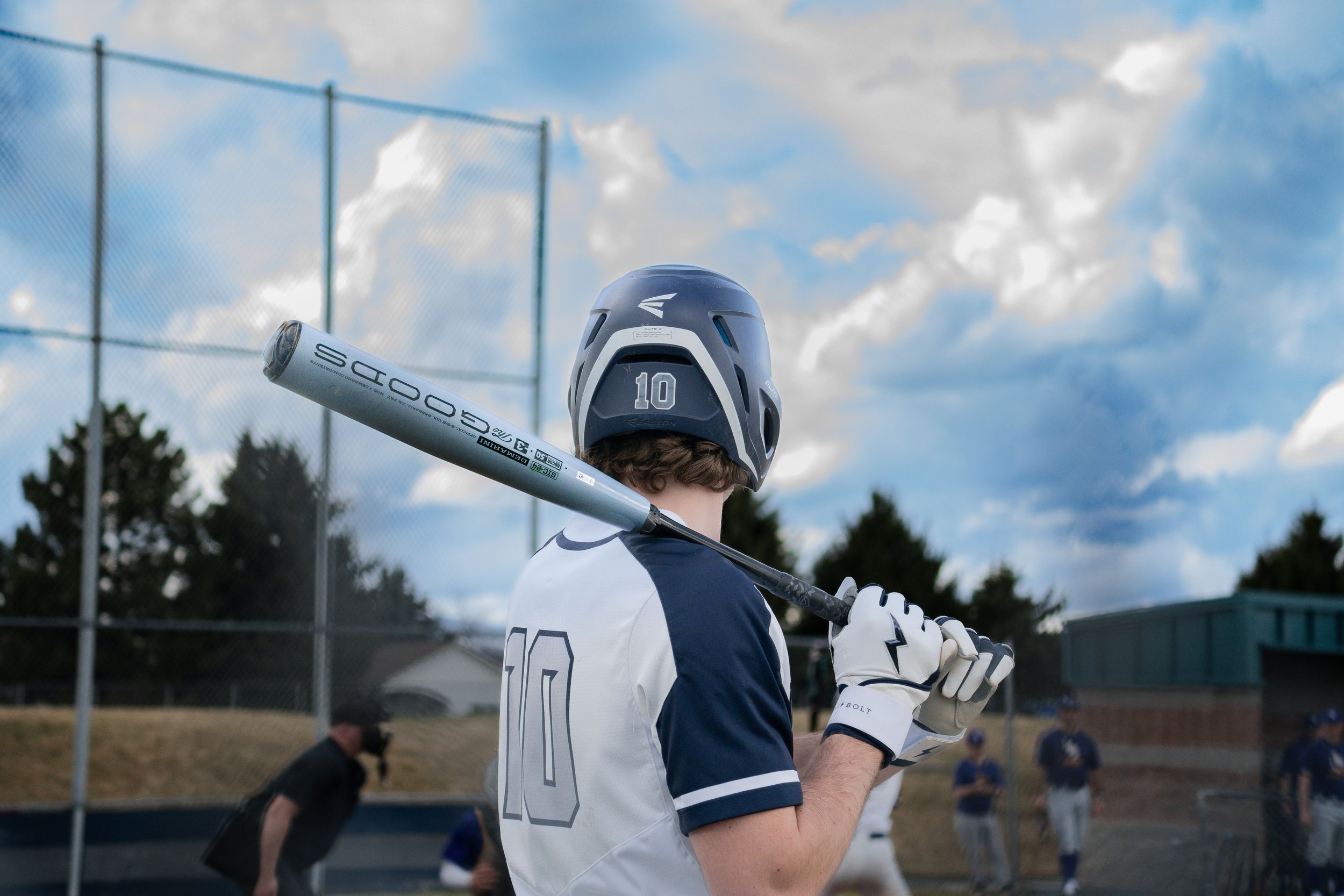 Baseball player in uniform holding a bat on shoulder at a field, wearing helmet and gloves, with teammates in the background.