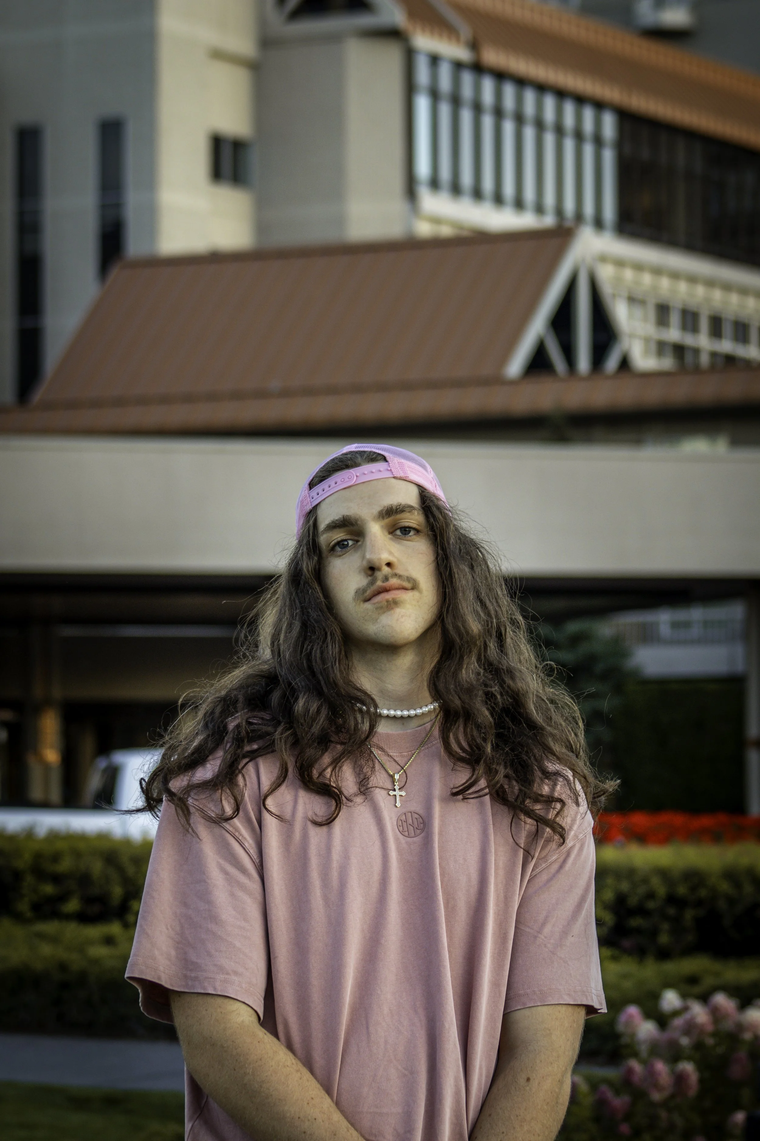 Young person with long hair wearing a pink shirt and cap, standing outdoors in front of a building.