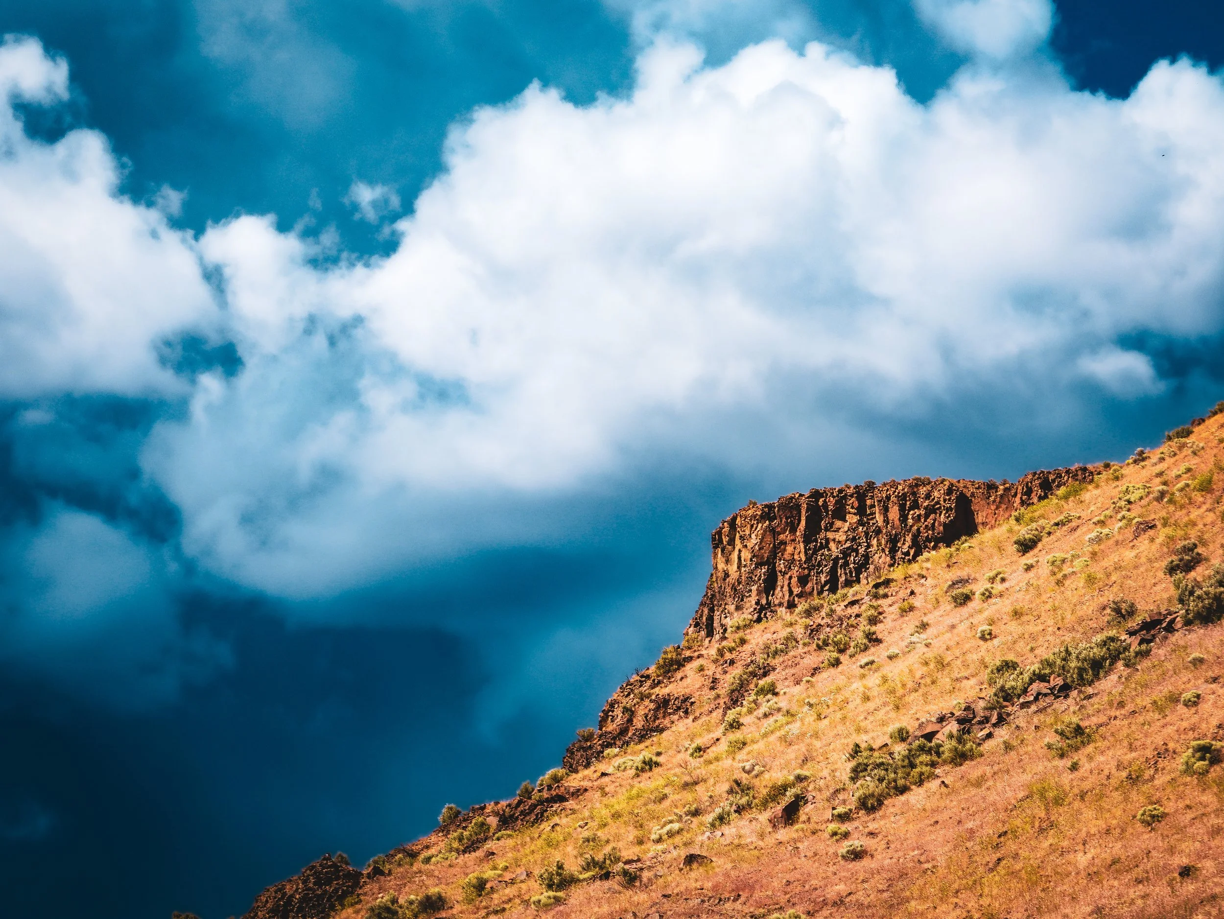 A scenic landscape featuring a hillside with sparse vegetation and rocky formations, with a partly cloudy blue sky overhead.