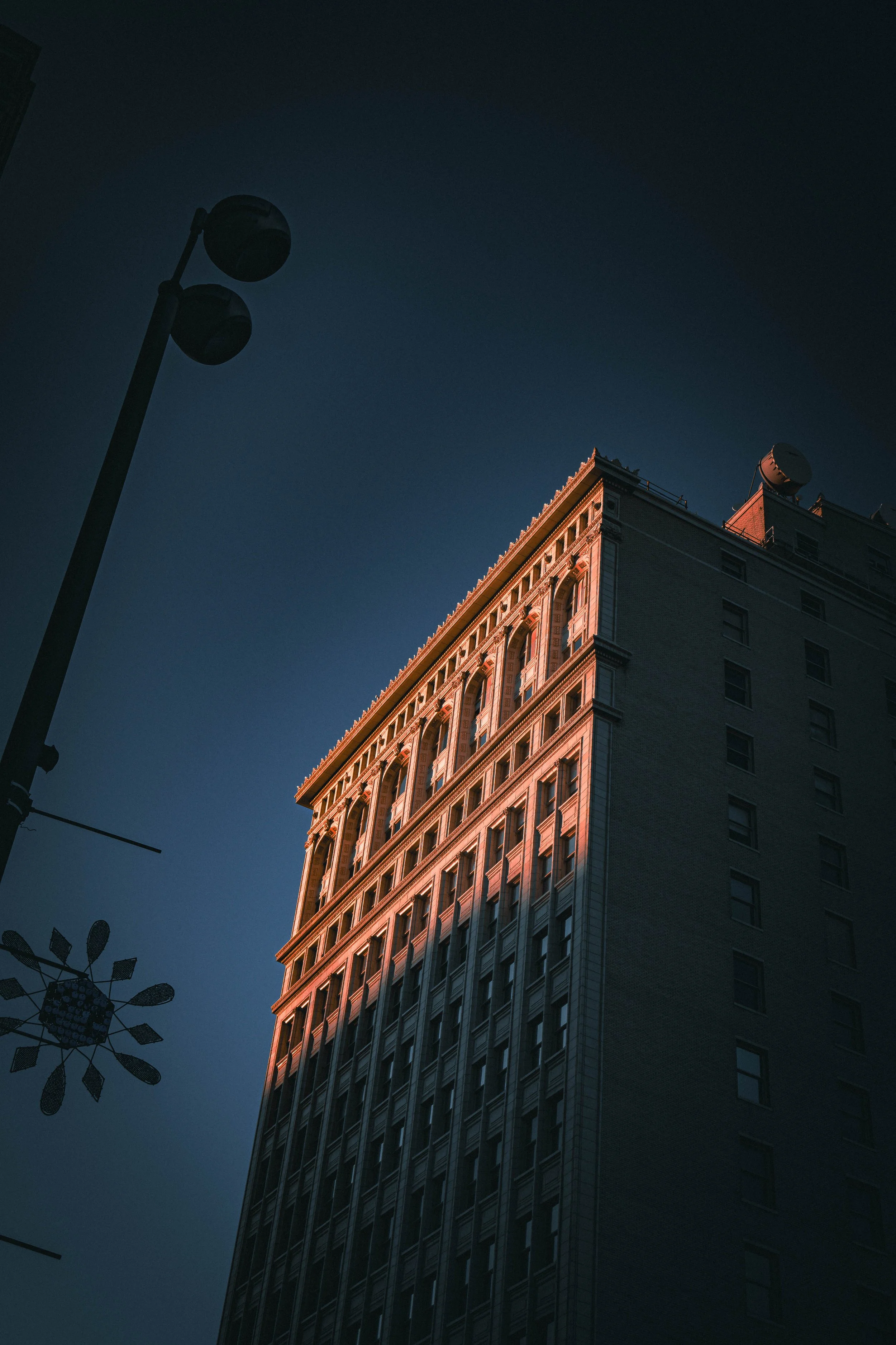 A tall city building during sunset with sunlight illuminating its upper floors, dark sky, and a streetlamp in the foreground.
