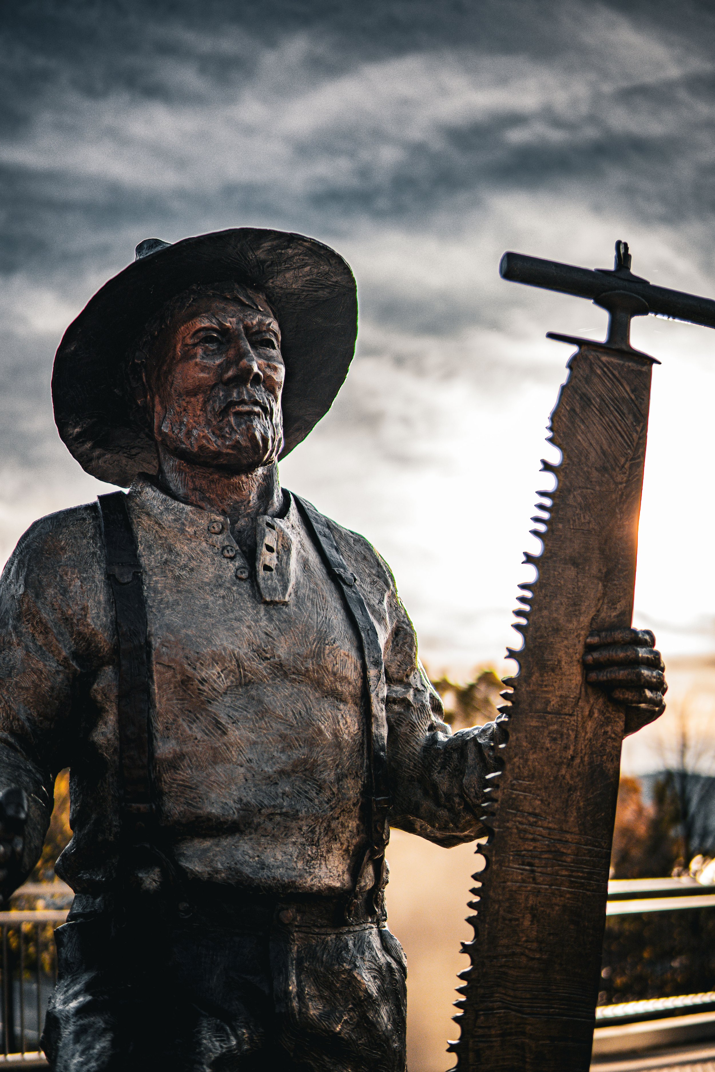 Bronze statue of a bearded man wearing a hat and holding a large saw, with a dramatic sky in the background.