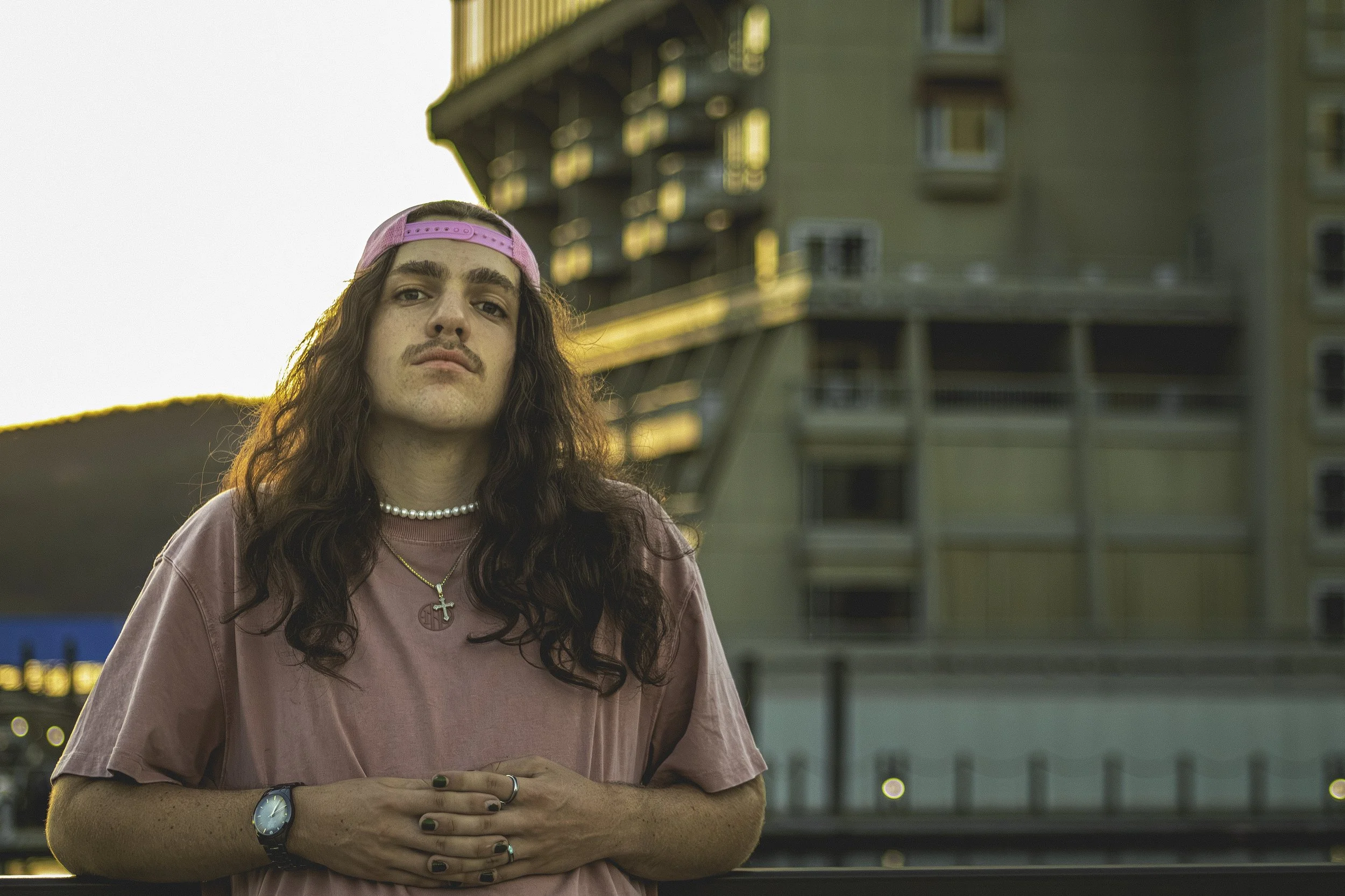 Person with long hair wearing a pink cap and shirt, standing in front of a building at sunset.