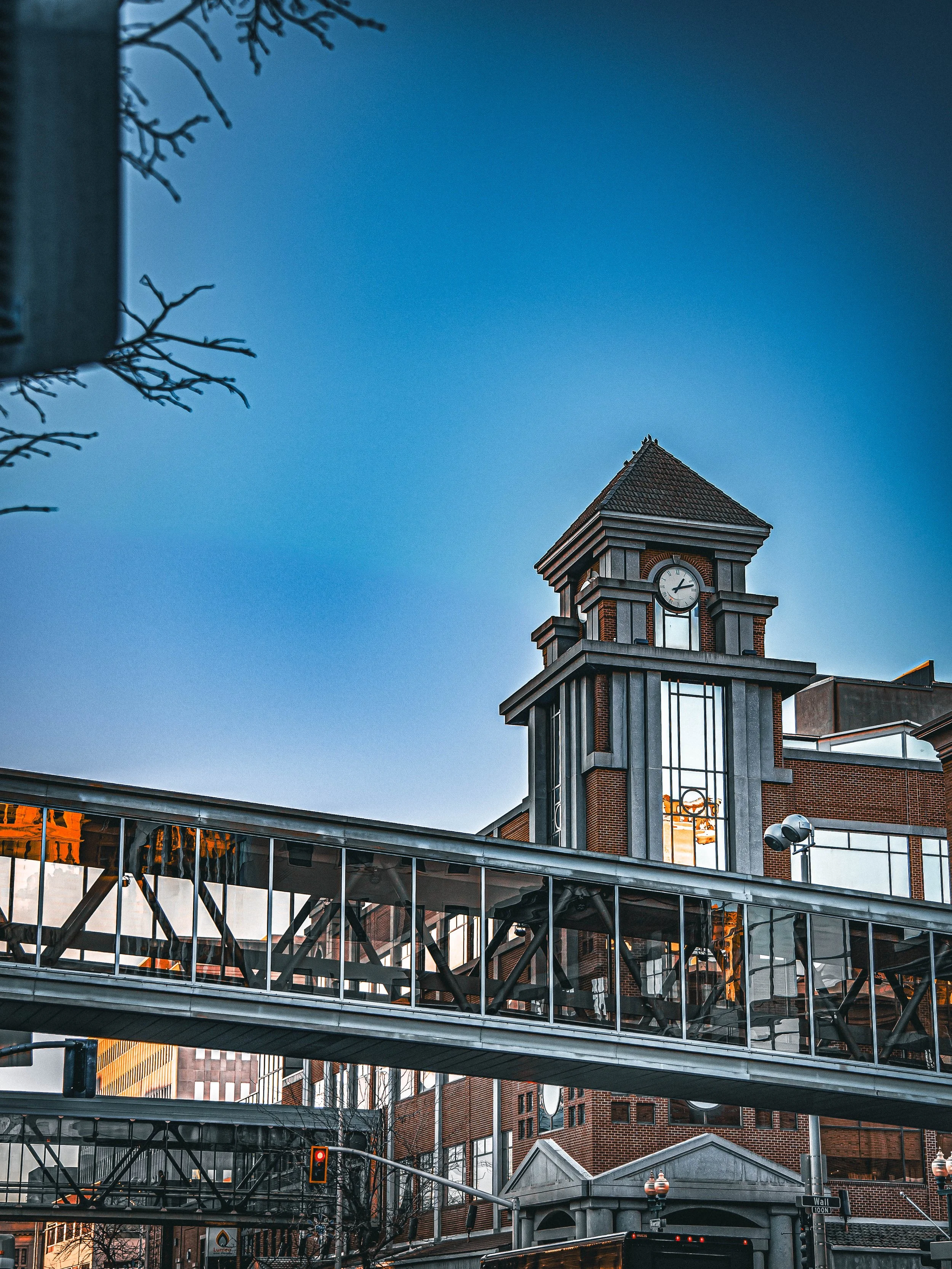 A tall brick building with a clock tower and glass-enclosed walkways in an urban setting, partially obscured by leafless tree branches, with an overcast sky.