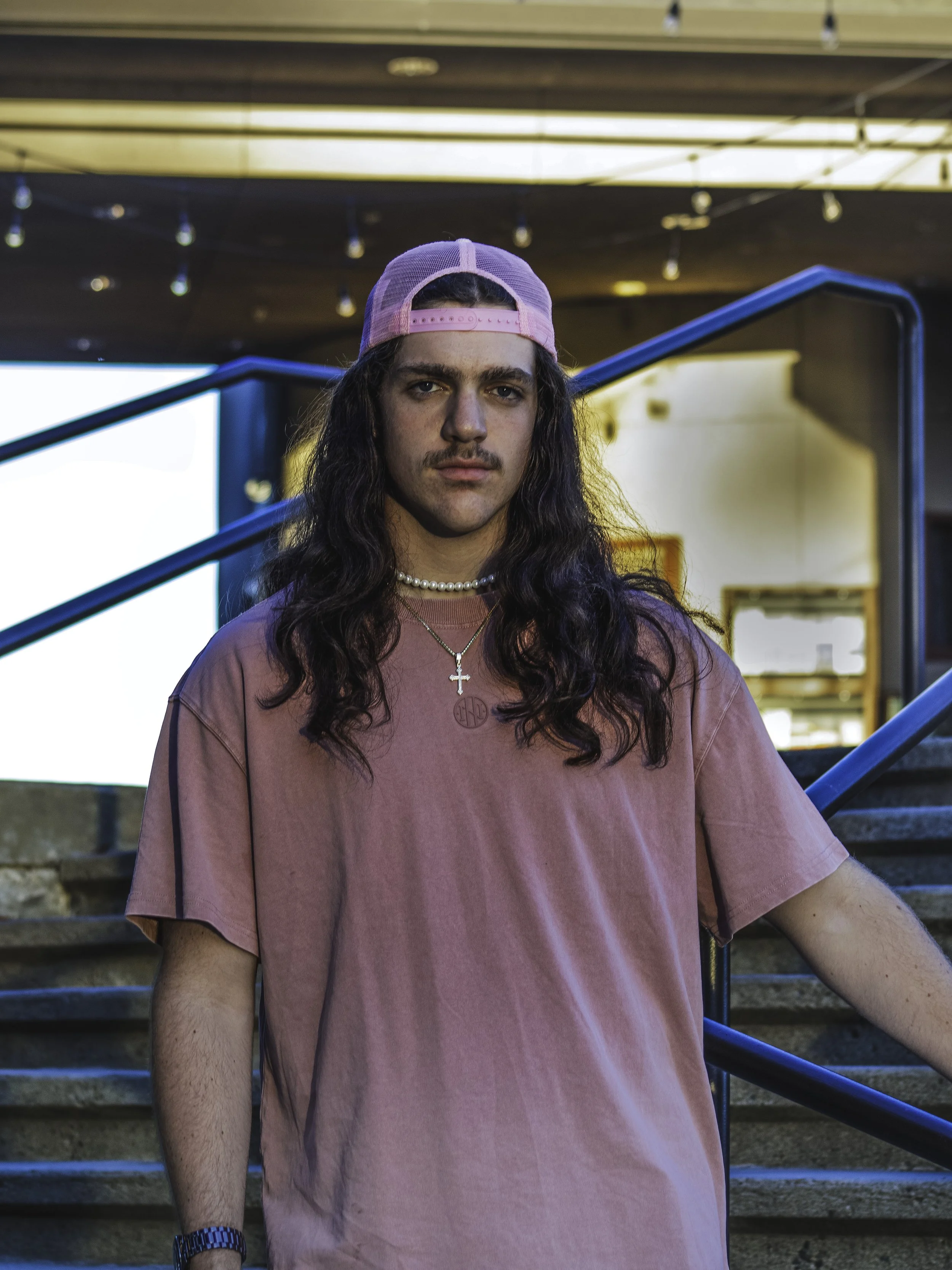Person with long hair wearing a pink hat and t-shirt, standing on stairs outdoors.