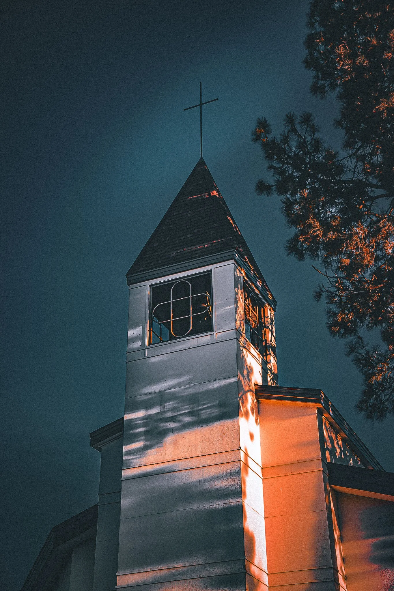 Church steeple with a cross at dusk, tree in foreground, illuminated by warm light.