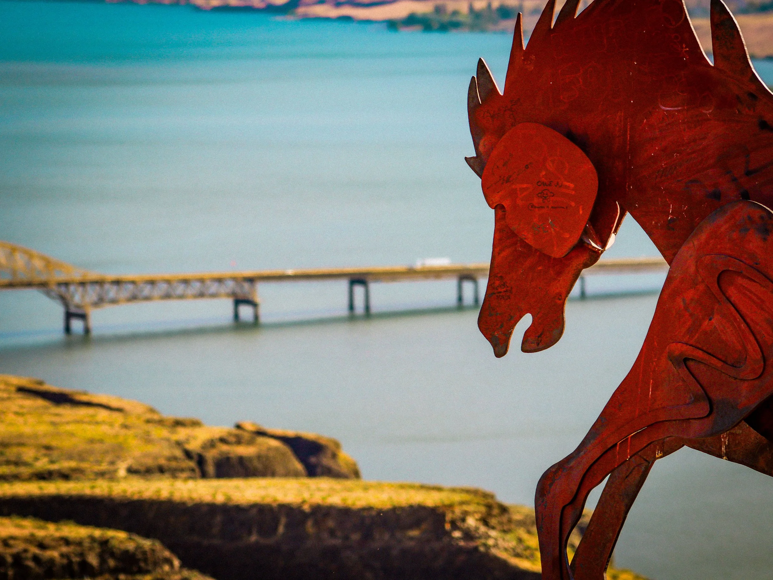 Close-up of a rusty metal sculpture of a horse's head during sunset near a body of water with a bridge in the background.