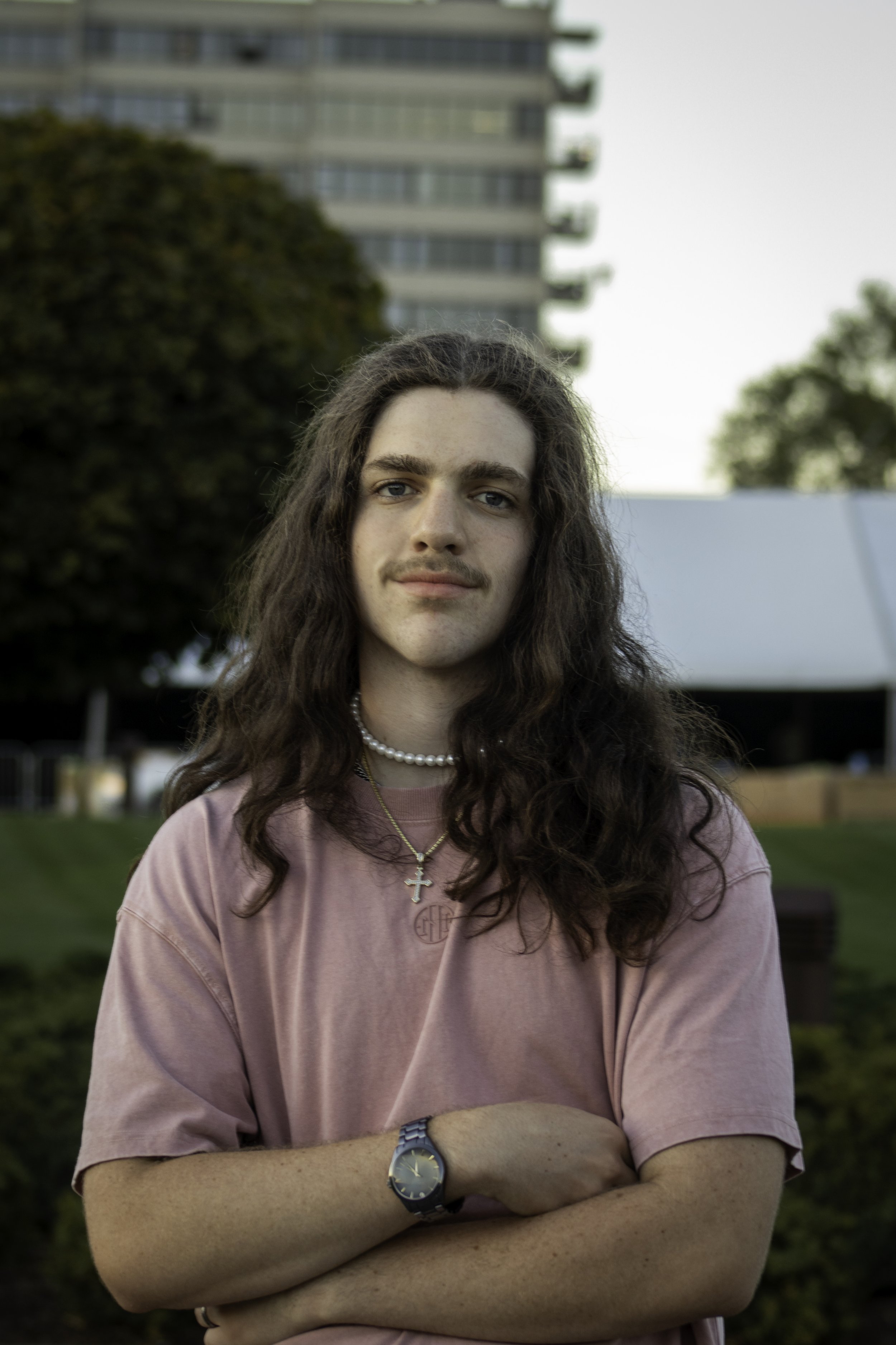 Person with long hair wearing a pink shirt, pearl necklace, and cross pendant, standing outside with trees and a building in the background.