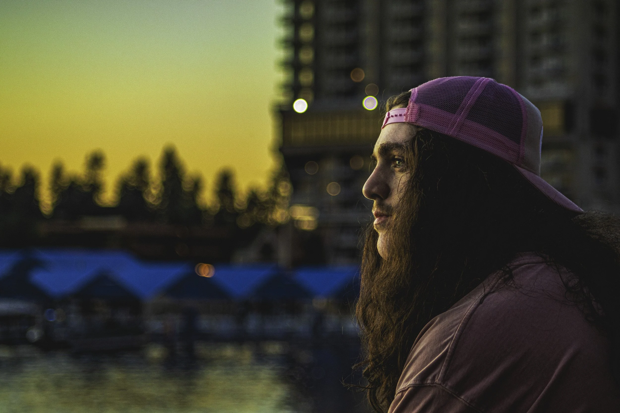 A woman with long hair and a beard, wearing a pink and black cap backwards, looks to the right during sunset near a body of water with blurry buildings and trees in the background.