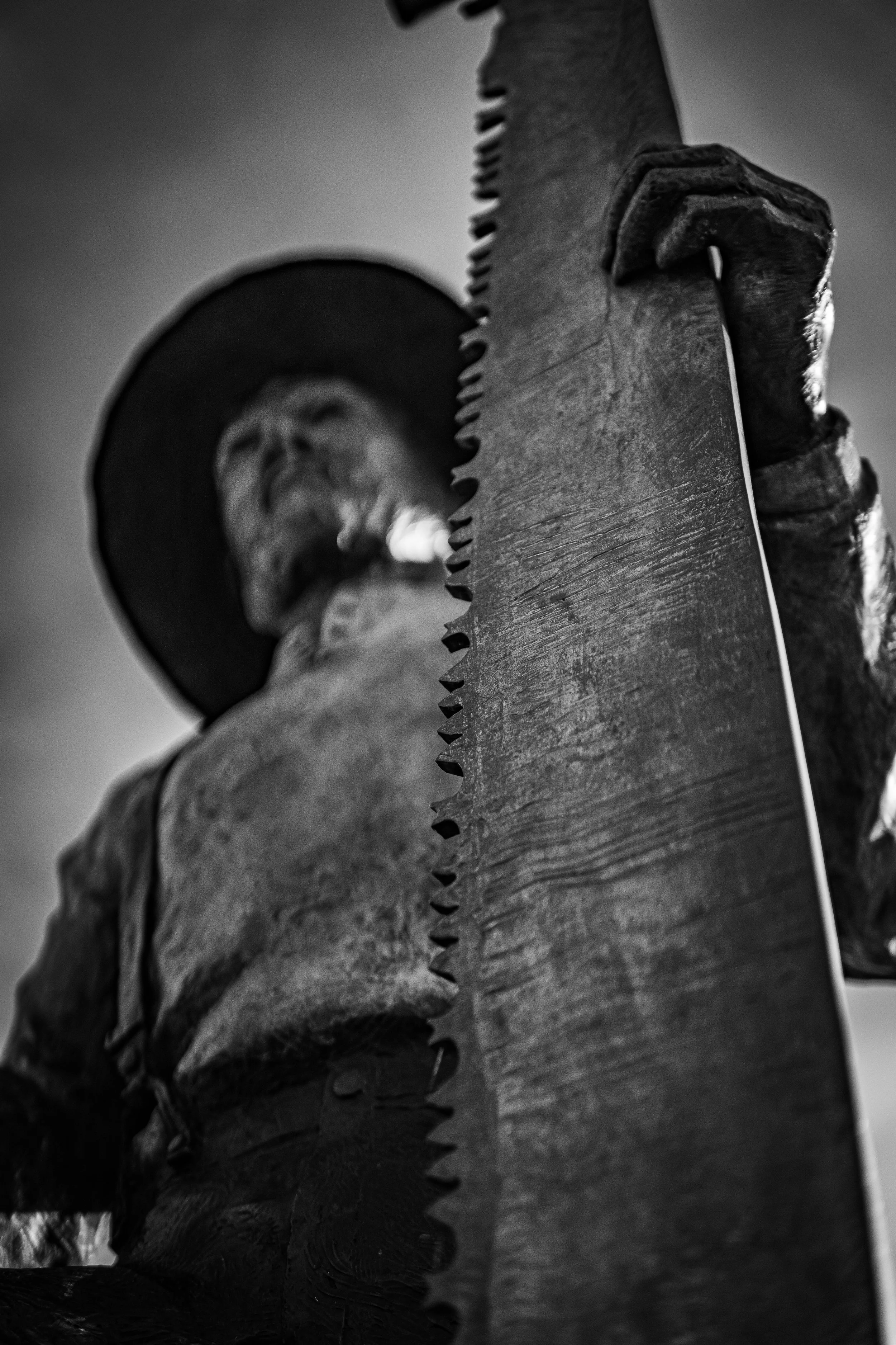 Close-up black and white image of a statue depicting a man with a hat holding a large saw, focusing on the saw teeth.