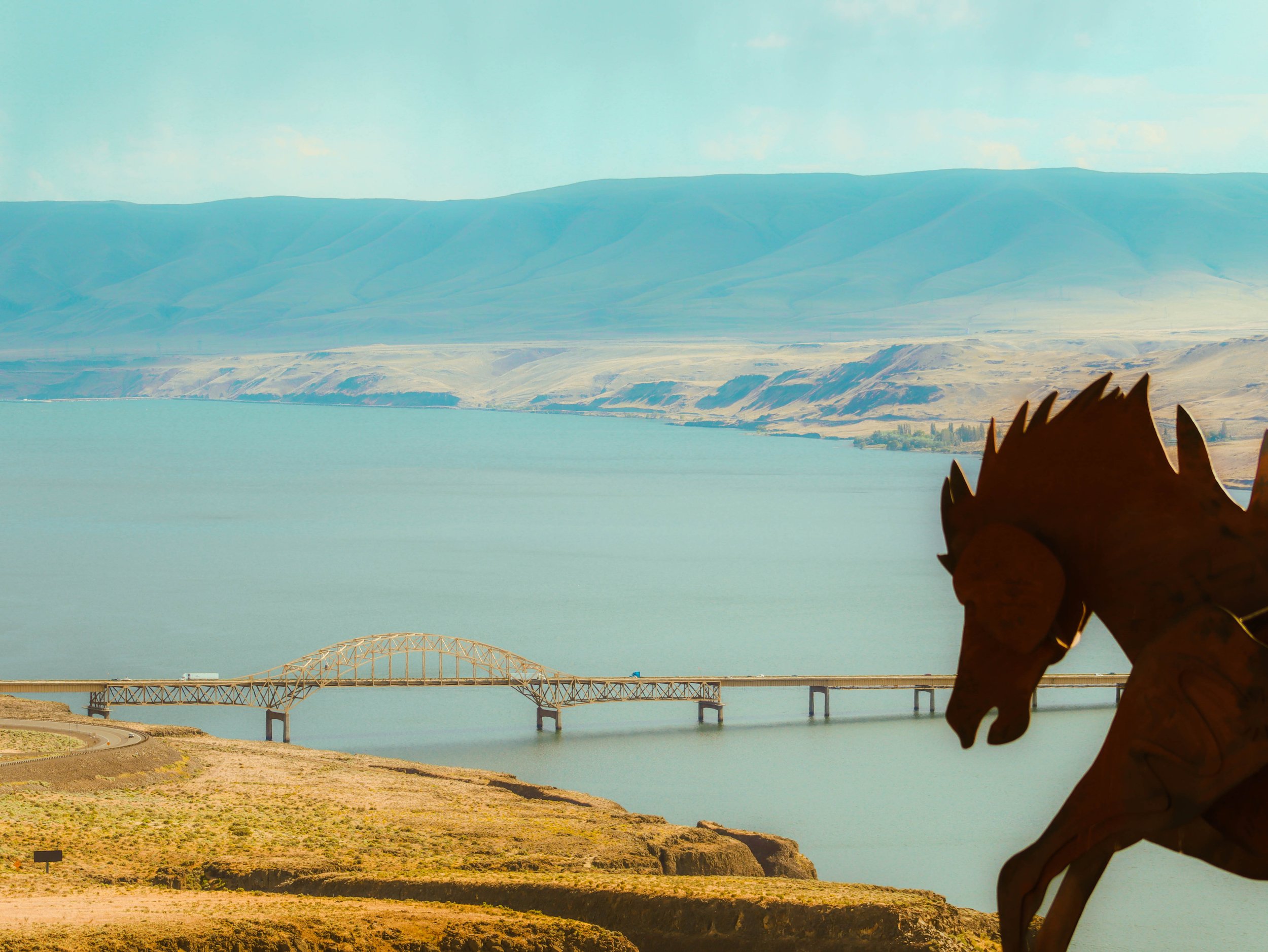 Silhouette of a horse sculpture in the foreground with a landscape of a bridge over a body of water, hills, and mountains in the background.