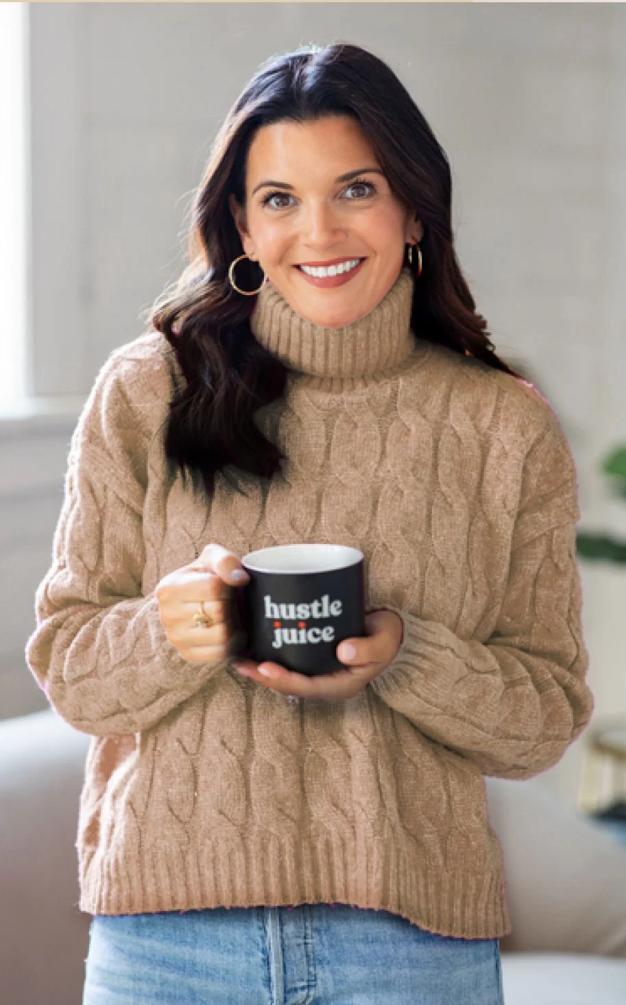Woman smiling with dark hair, wearing a beige turtleneck sweater, holding a black mug with "hustle juice" printed on it.