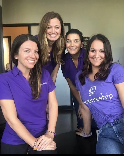 Four women smiling, wearing purple "Aspireship" shirts, standing indoors.