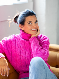 A woman in a bright pink sweater sitting on a couch with a cheerful expression, resting her chin on her hand.