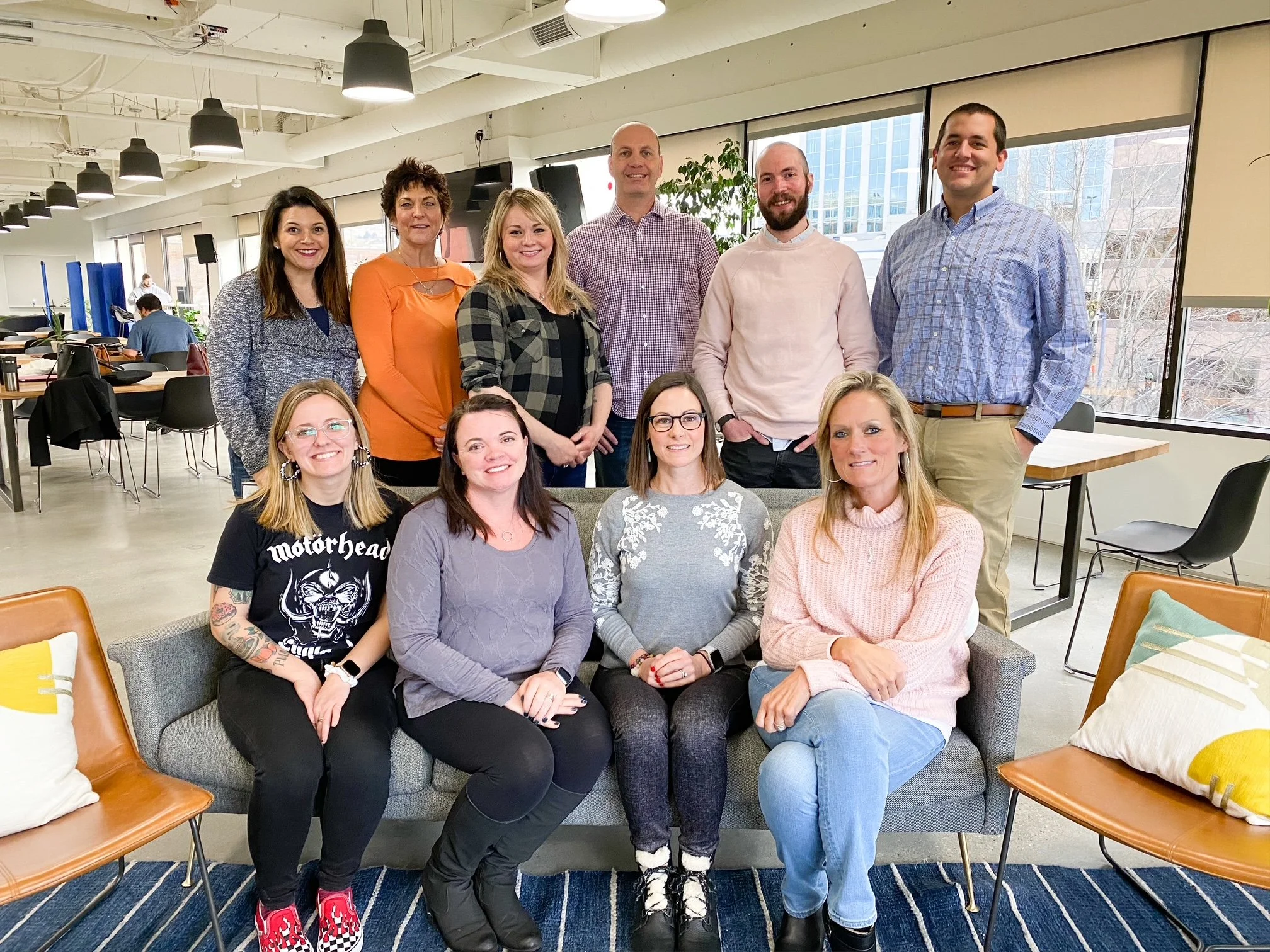 Group of ten people posing indoors in a modern office space. They are seated and standing in two rows, smiling at the camera.