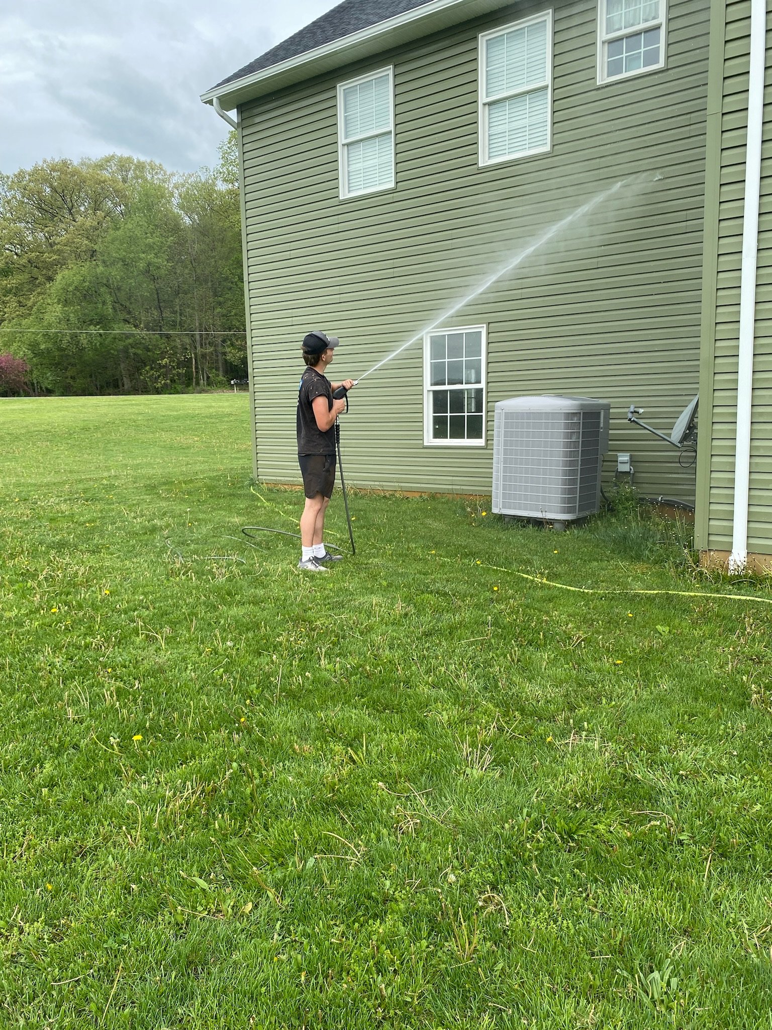 A person wearing a baseball cap, headphones, a black t-shirt, shorts, and sneakers is watering the exterior wall of a green house with a garden hose.