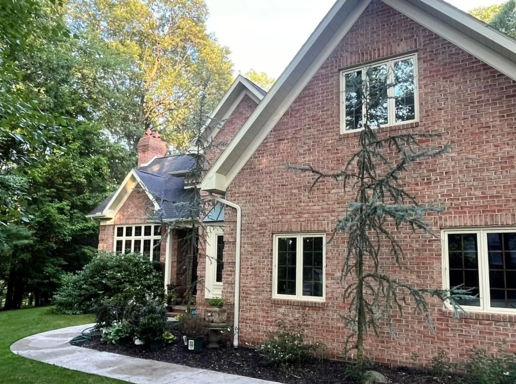 A brick house with multiple windows, surrounded by trees and a well-maintained lawn. There's a curved sidewalk in front and a front porch partially visible on the left.