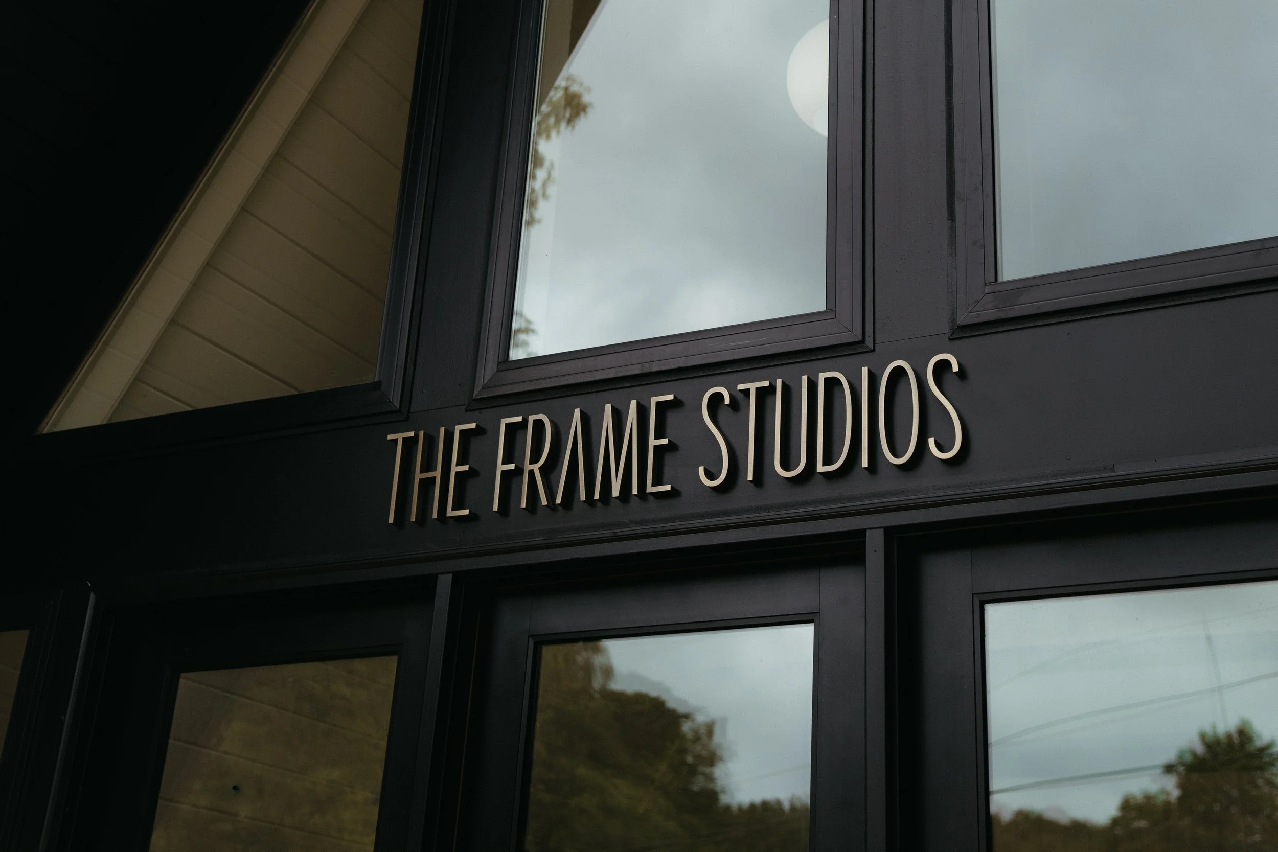 Close-up of a building storefront with a black sign reading 'THE FRAME STUDIOS' in beige letters, reflecting the sky and trees in the glass windows above.