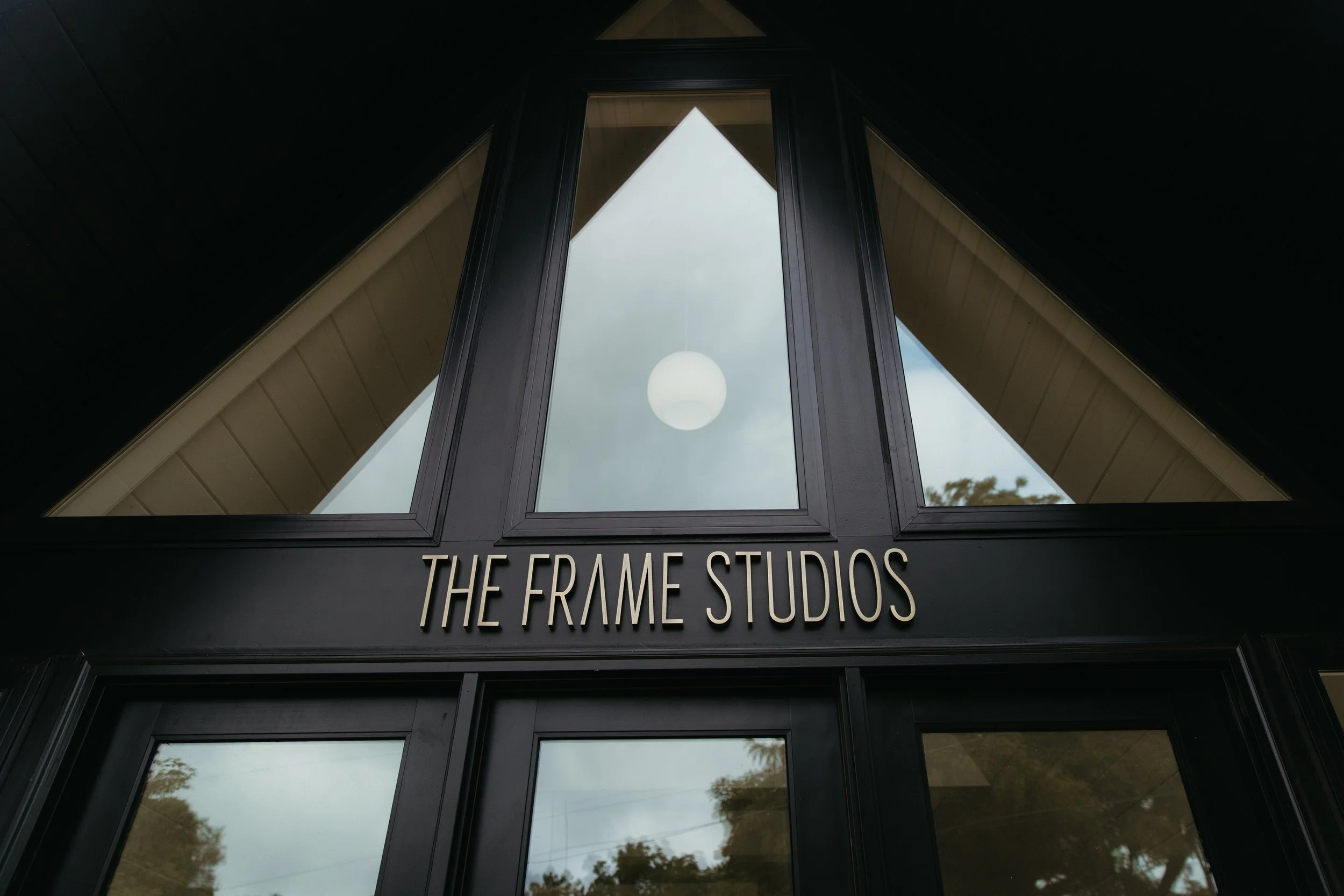 Close-up view of the entrance to The Frame Studios, with large triangular window and black framing, reflecting the sky and trees.