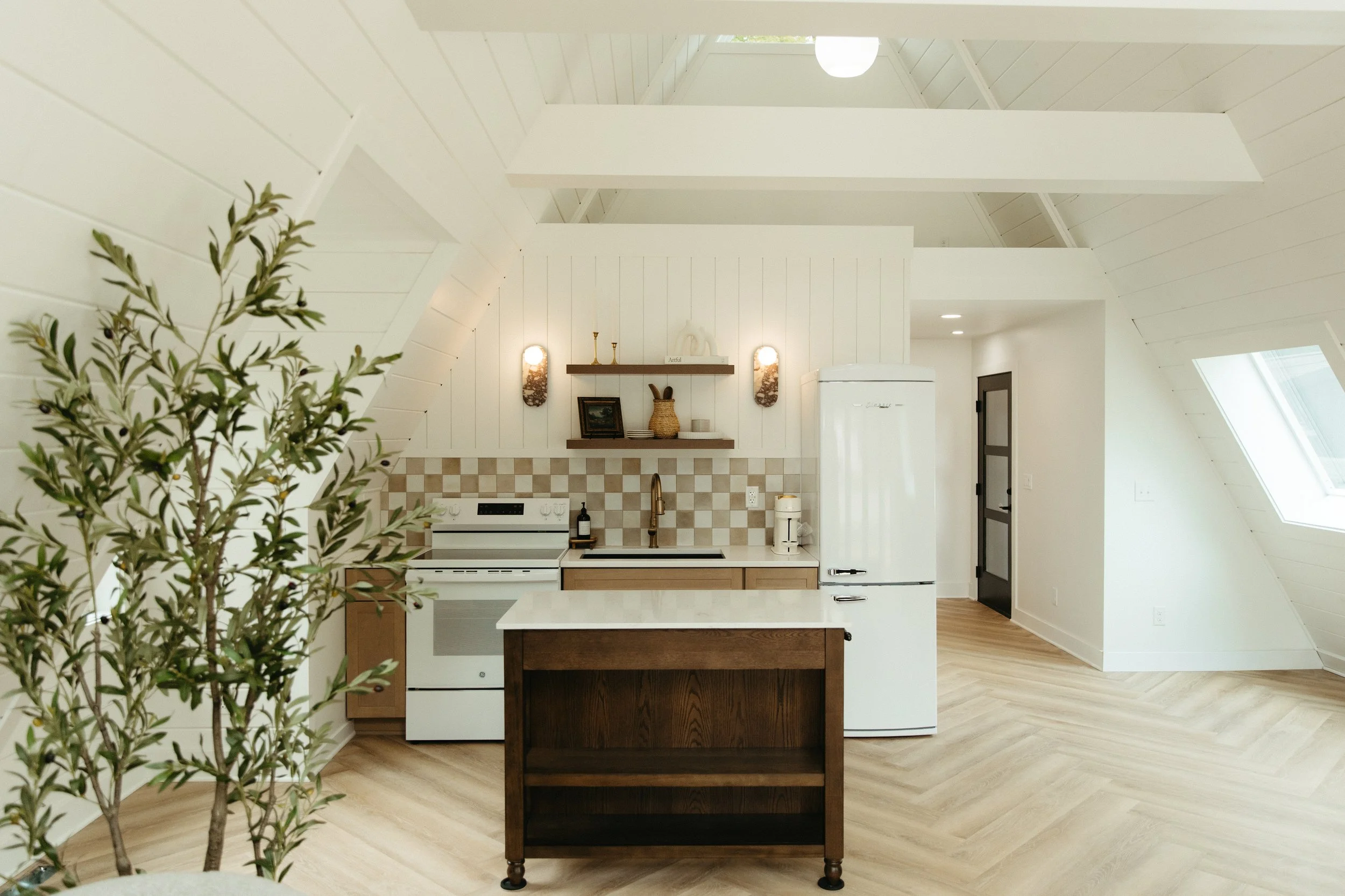 A white kitchen with a small kitchen island, a white refrigerator, and a black door. The ceiling is high with white wooden beams and skylight windows, and there's a small potted plant in the foreground.