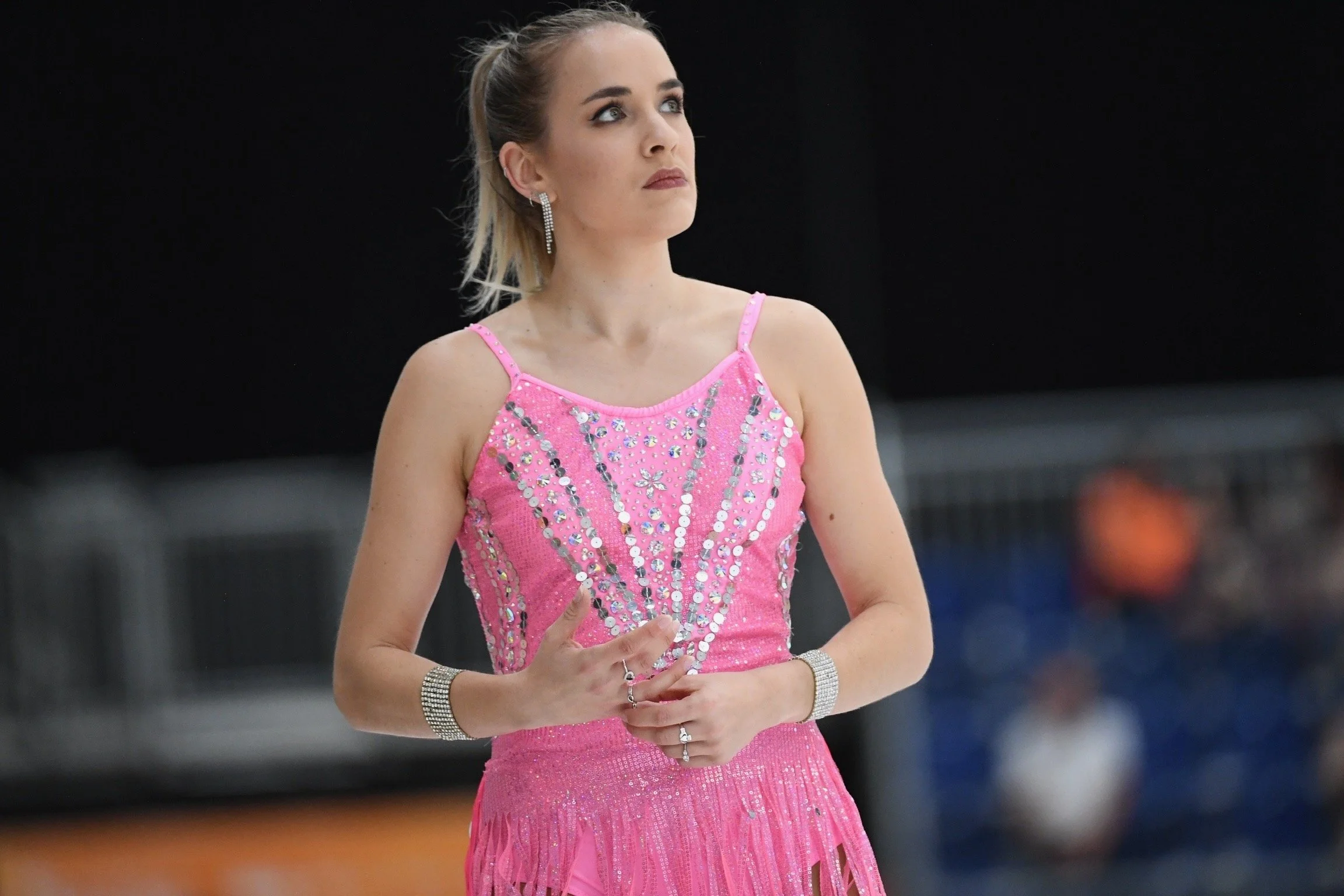 Female figure skater in a pink dress adorned with rhinestones, standing on ice with her hands clasped in front of her.