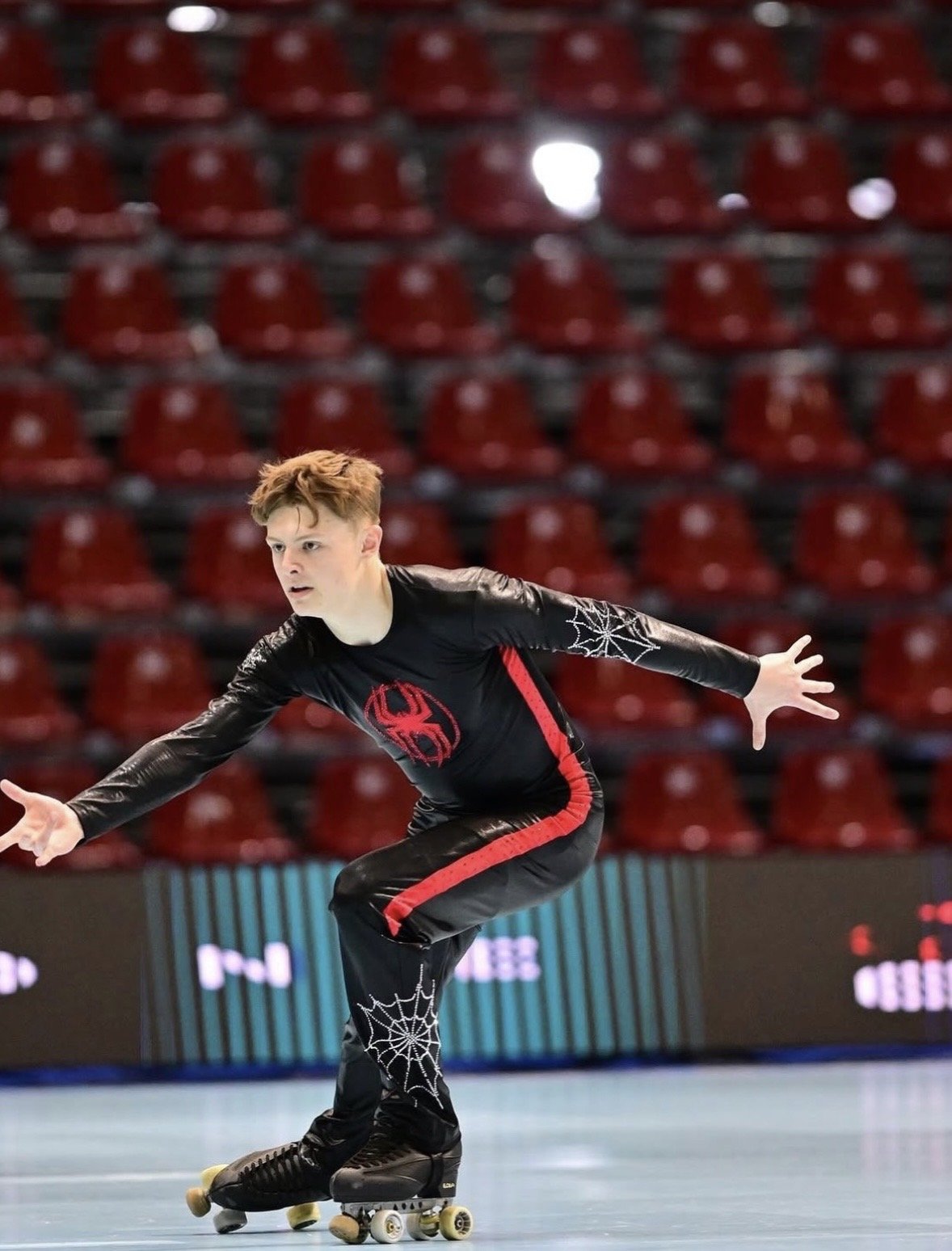 A young male roller skater in a black and red costume with spider web designs, performing on a roller rink with red seating in the background.