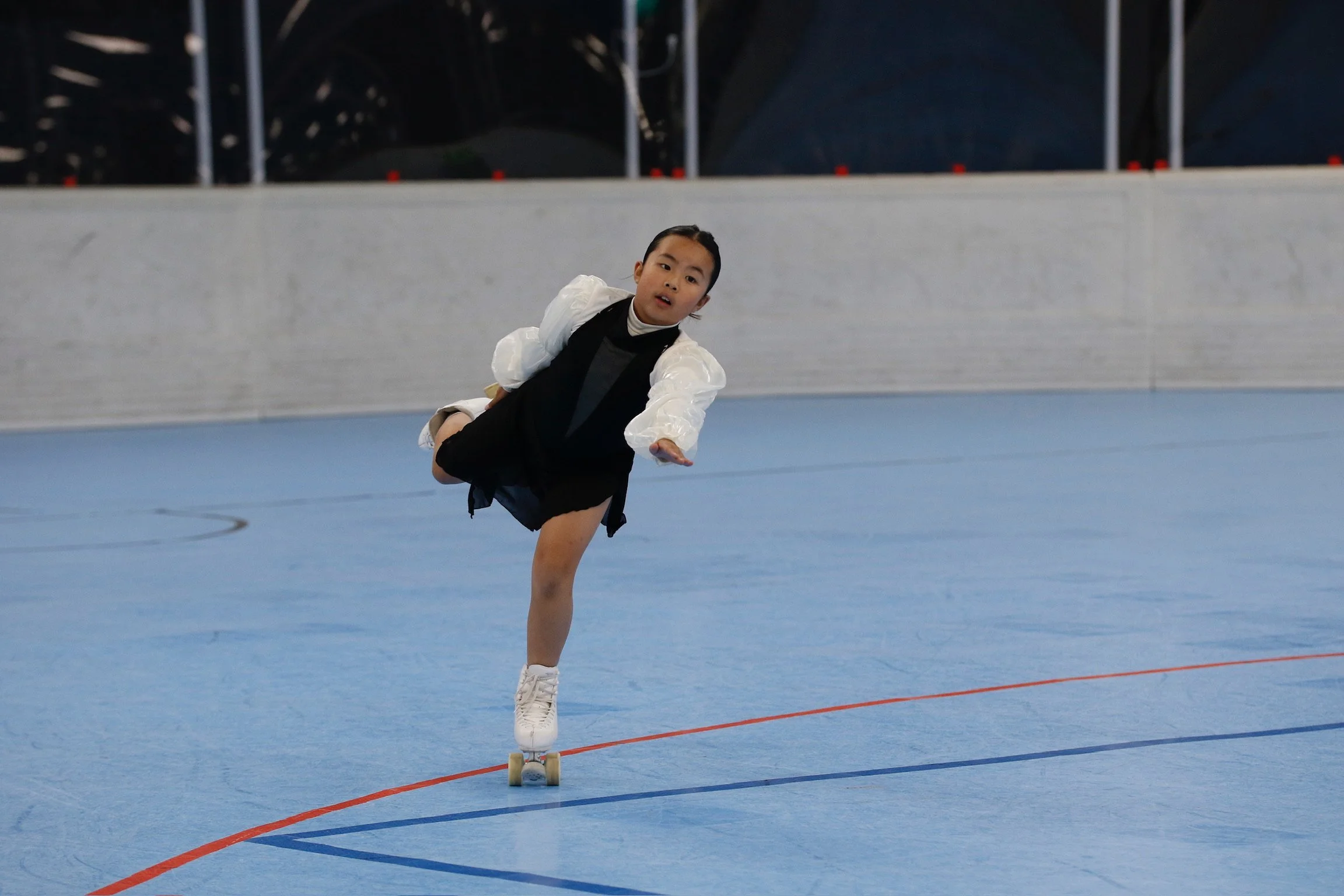 A young figure skater in black and white outfit performing a move on the ice rink. The skater is balancing on one leg with the other leg extended back. The background shows the ice rink and a dark indoor setting.
