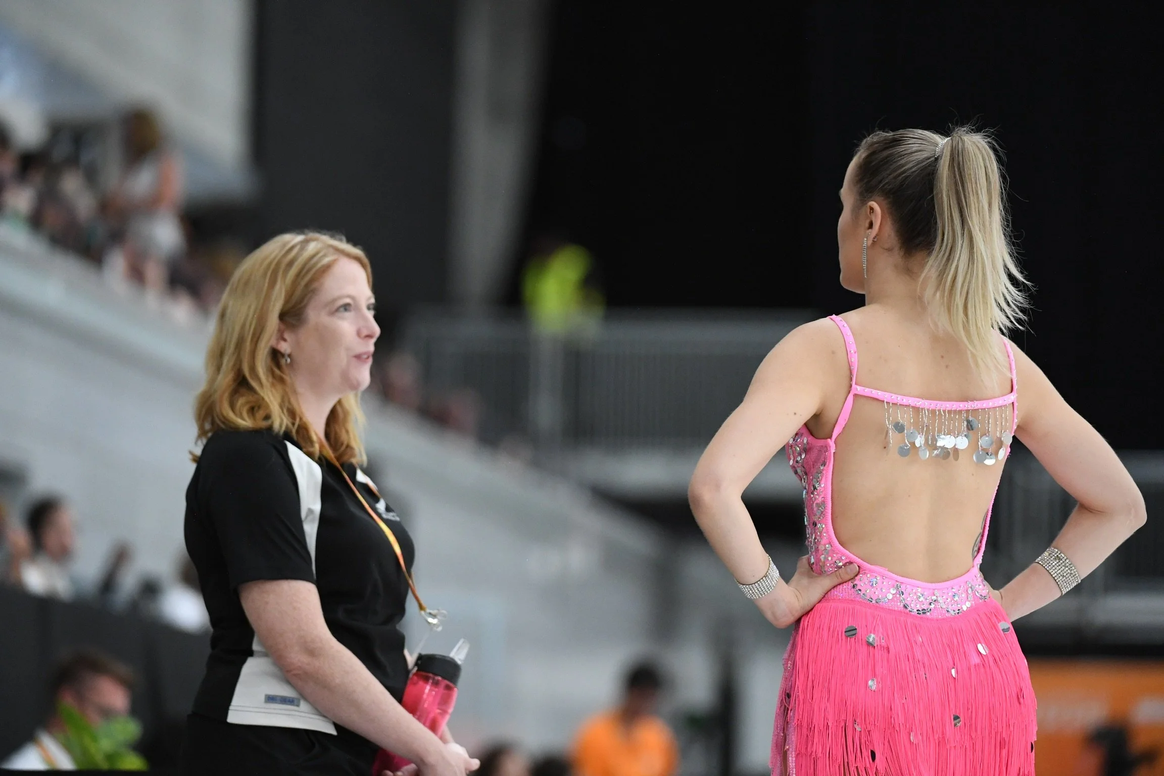 A female figure skater in a pink costume with silver embellishments talking to a woman in black and white clothing at an indoor skating rink.