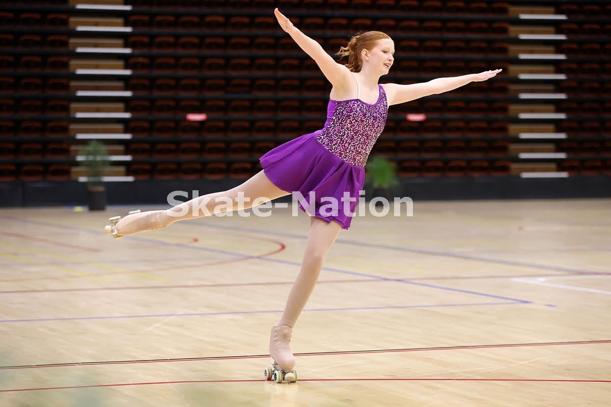 A young female figure skater in a purple dress performing a move on roller skates in an indoor skating rink.