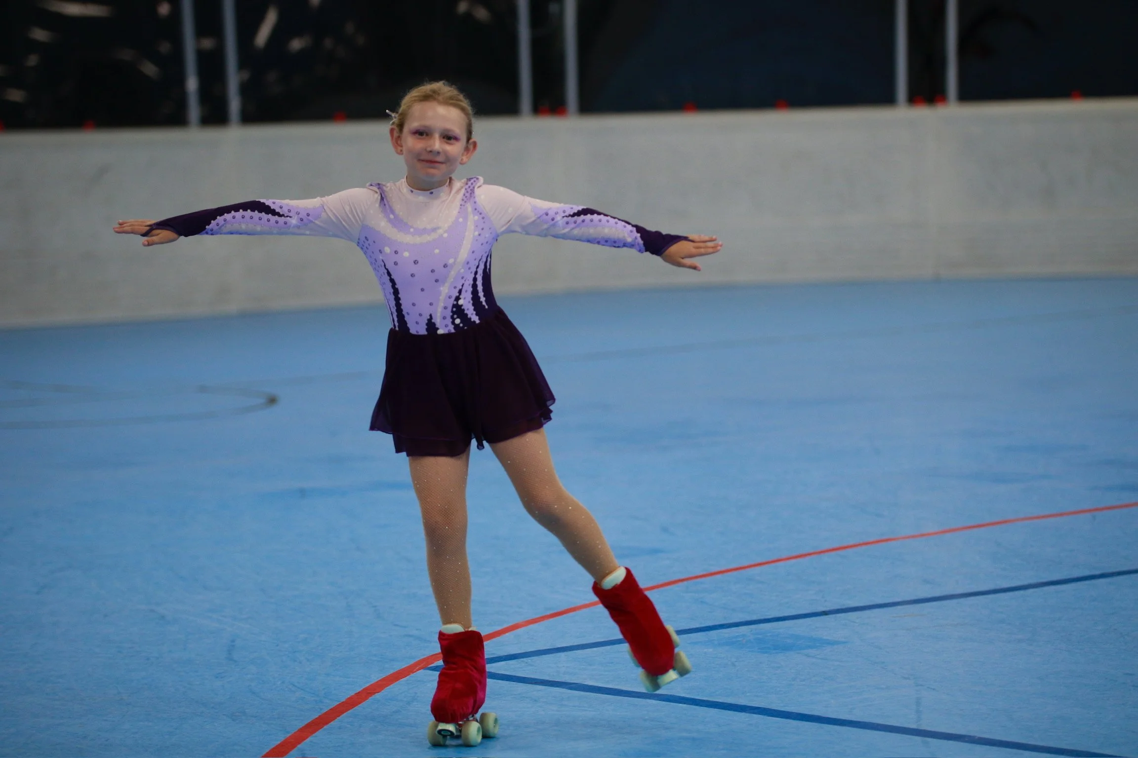 young female roller skater performing a move on a skating rink on 1 foot wearing a purple leotard