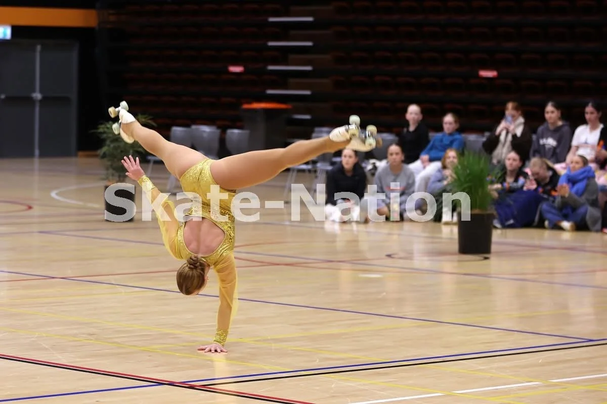 A female roller skater performing a handstand on a gymnasium floor during a skating event, with spectators seated in the background.