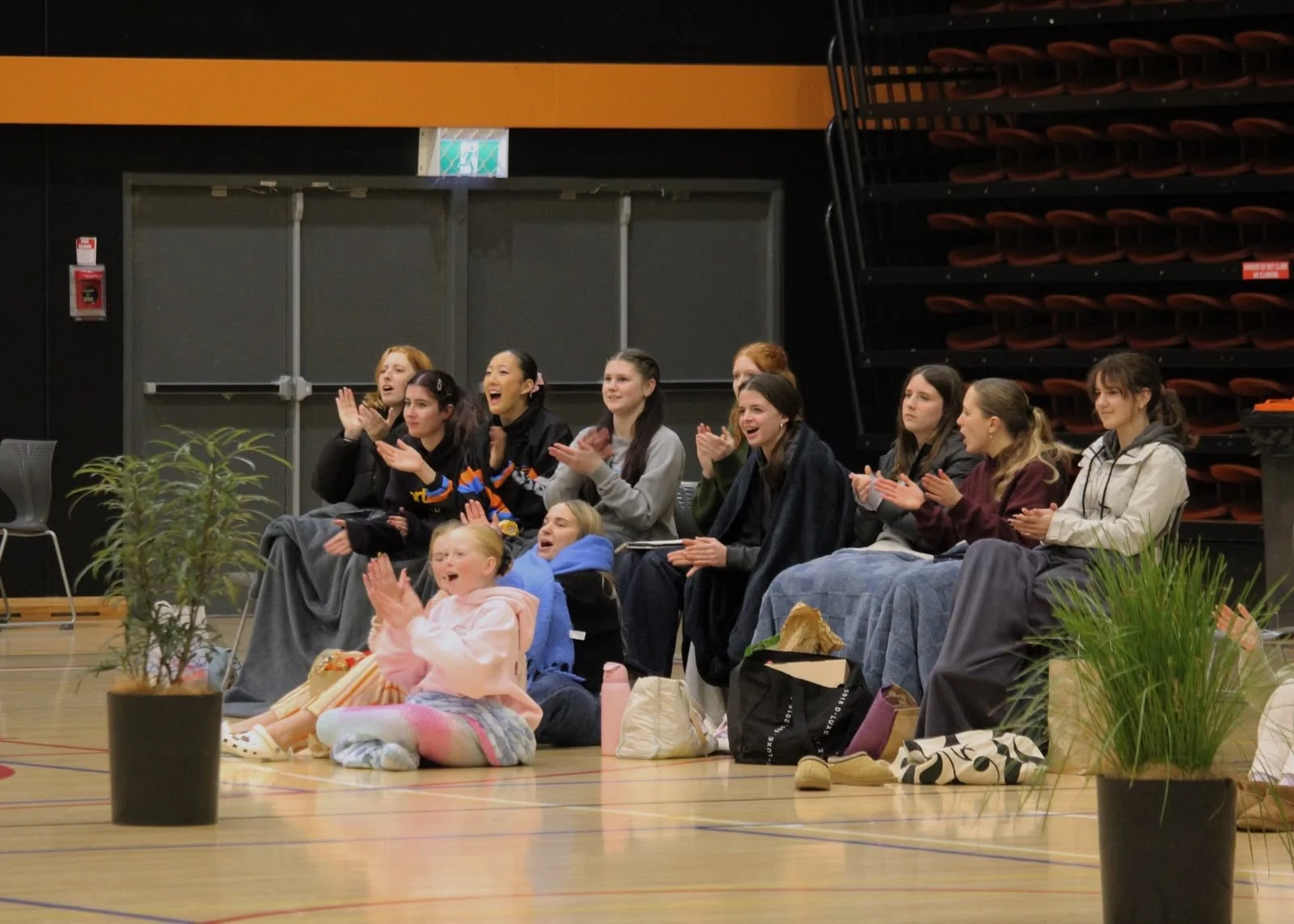 Group of young women and girls sitting on the floor and in chairs in a gymnasium, clapping and smiling during an event, with potted plants in the foreground.
