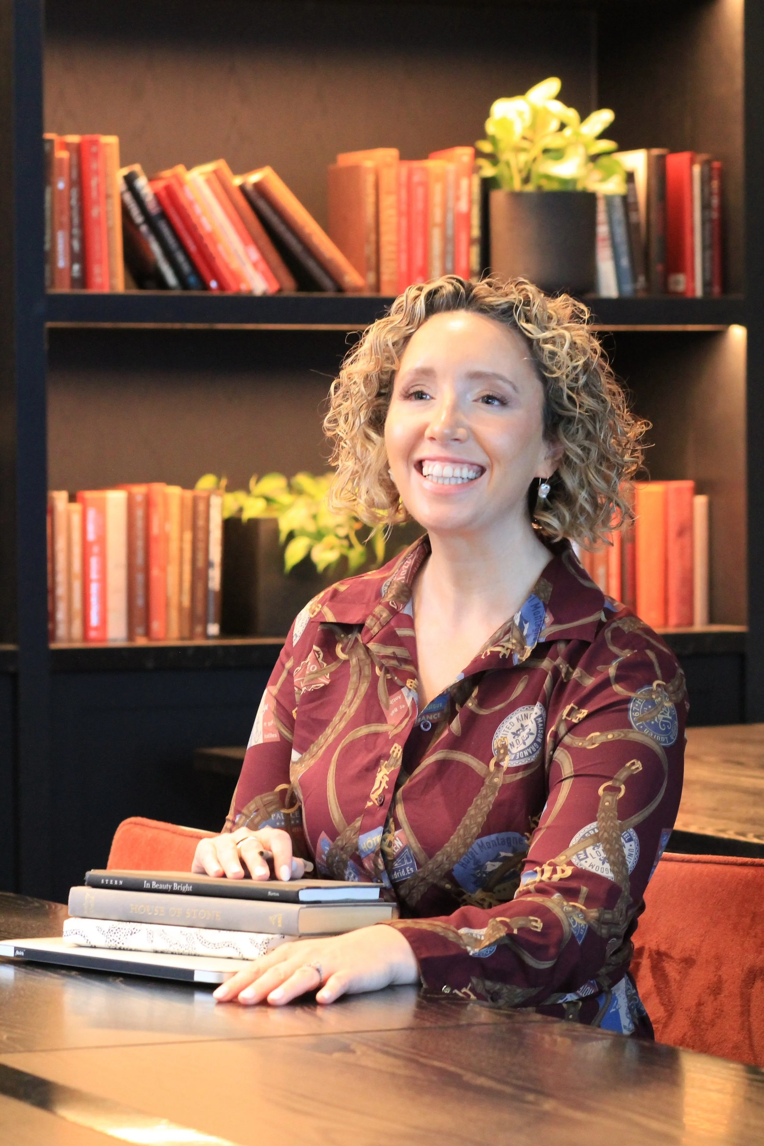 A woman with curly blonde hair smiling and sitting at a wooden table with books in front of her, in a room with black bookshelves filled with red and orange books and a potted plant.
