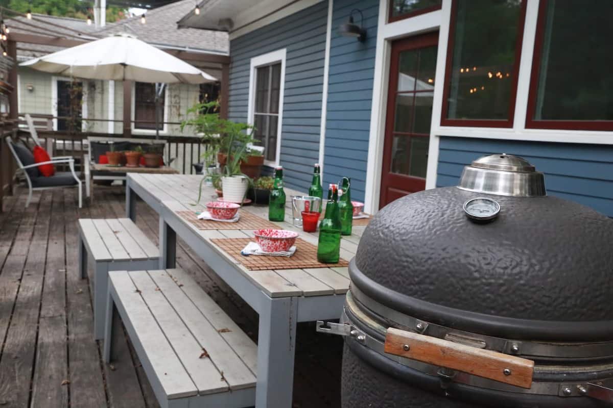 Backyard patio with a wooden table set with drinks and bowls, a charcoal barbecue grill in the foreground, and outdoor seating with a patio umbrella in the background.